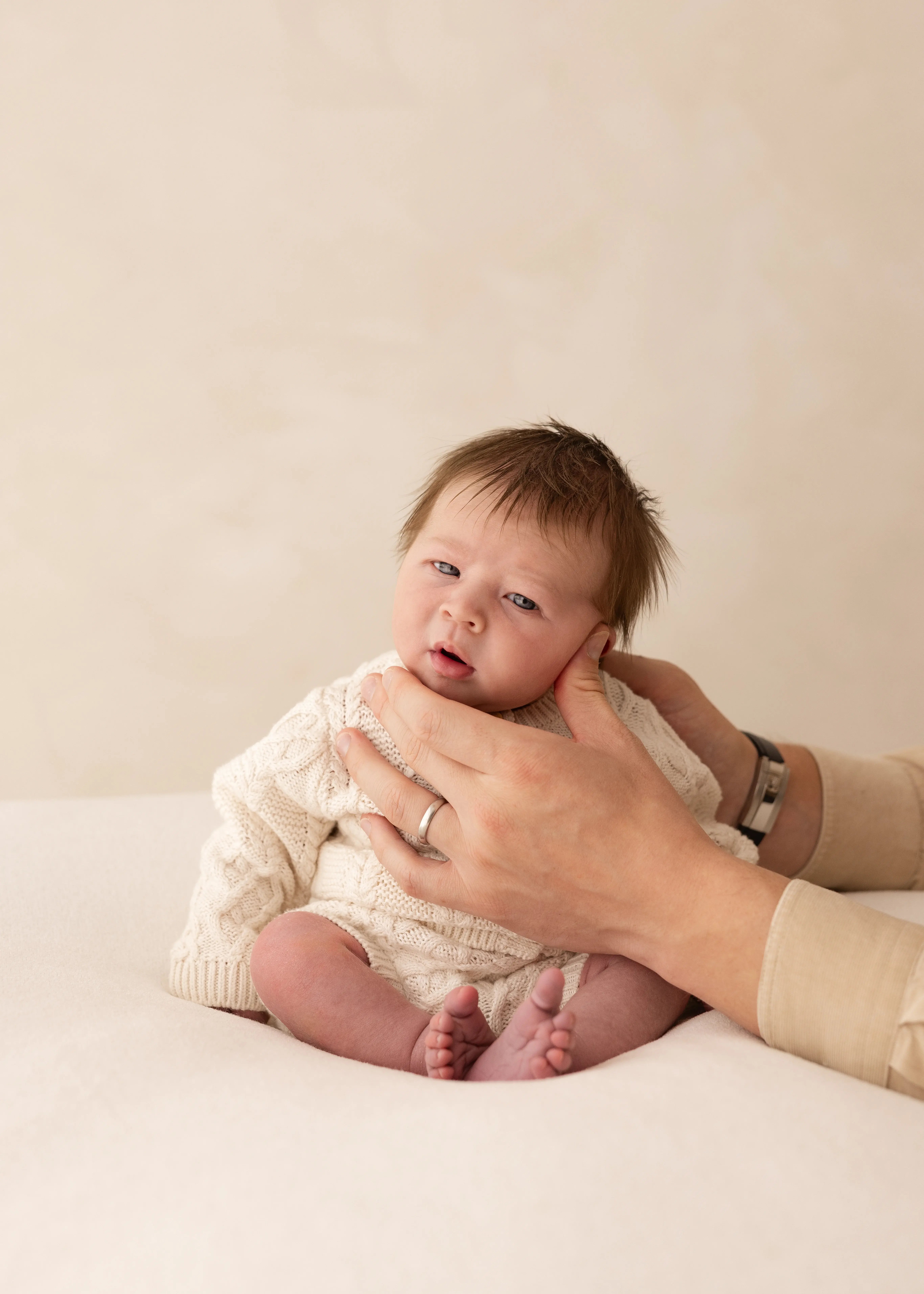 Newborn baby in a cream knitted outfit being gently supported, looking wide awake during a natural newborn photoshoot in Kent.