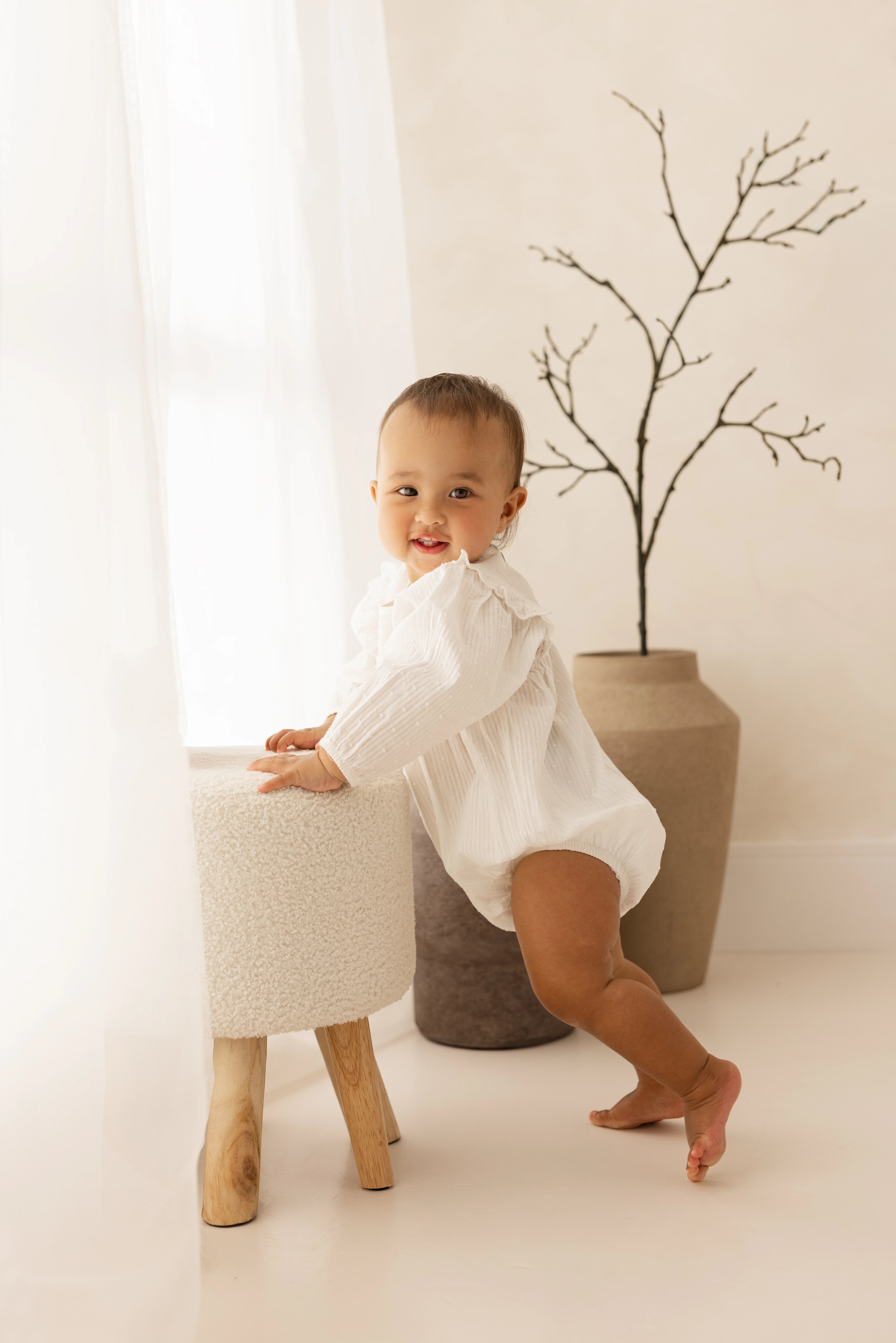 Smiling baby in a white long-sleeved romper stands holding a textured cream stool beside a window with soft natural light, with decorative vases and a twig arrangement in the background.