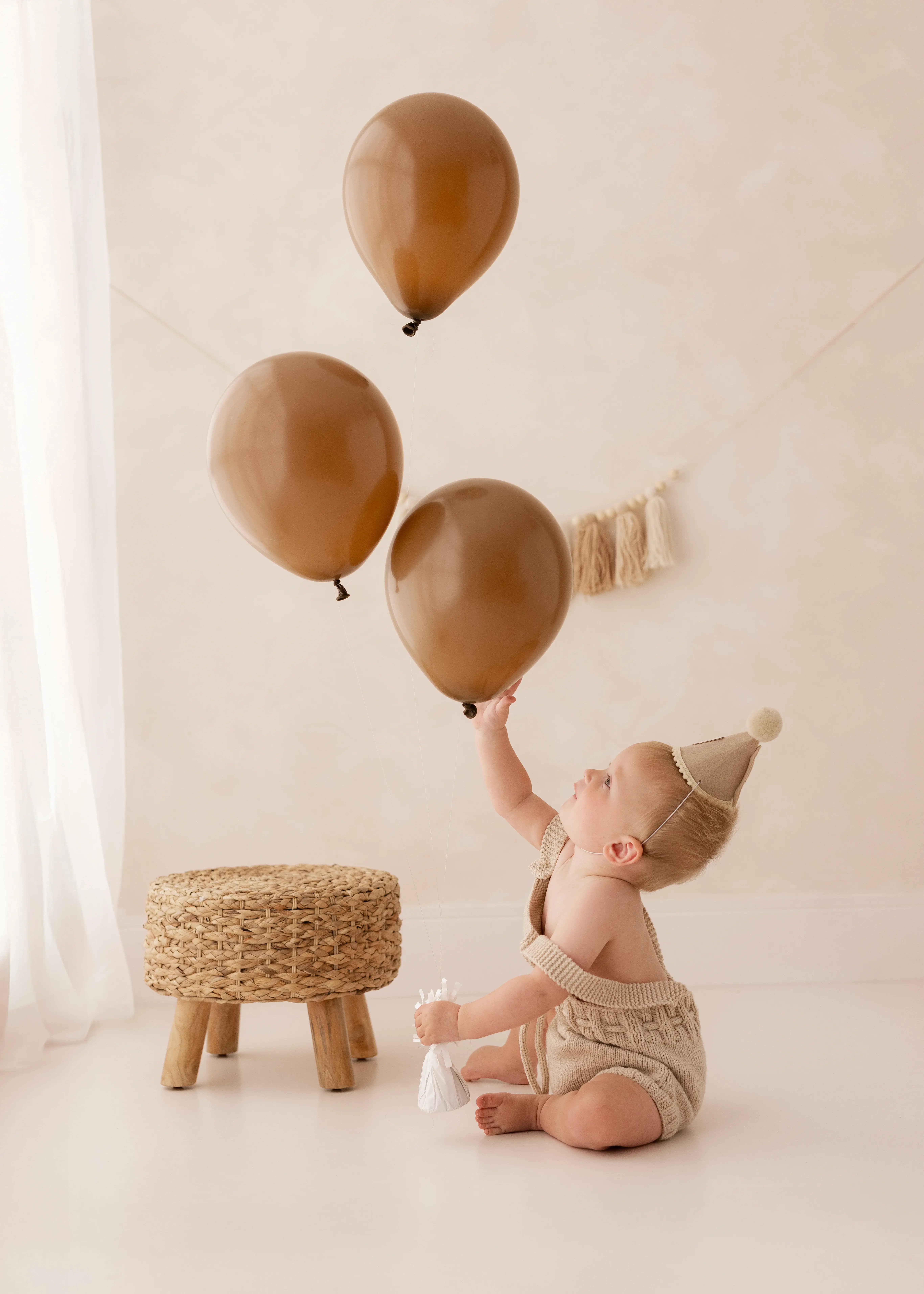 Baby wearing a first birthday party hat, sitting on the studio floor and reaching up toward a cluster of brown balloons during a natural first birthday photoshoot in Kent.