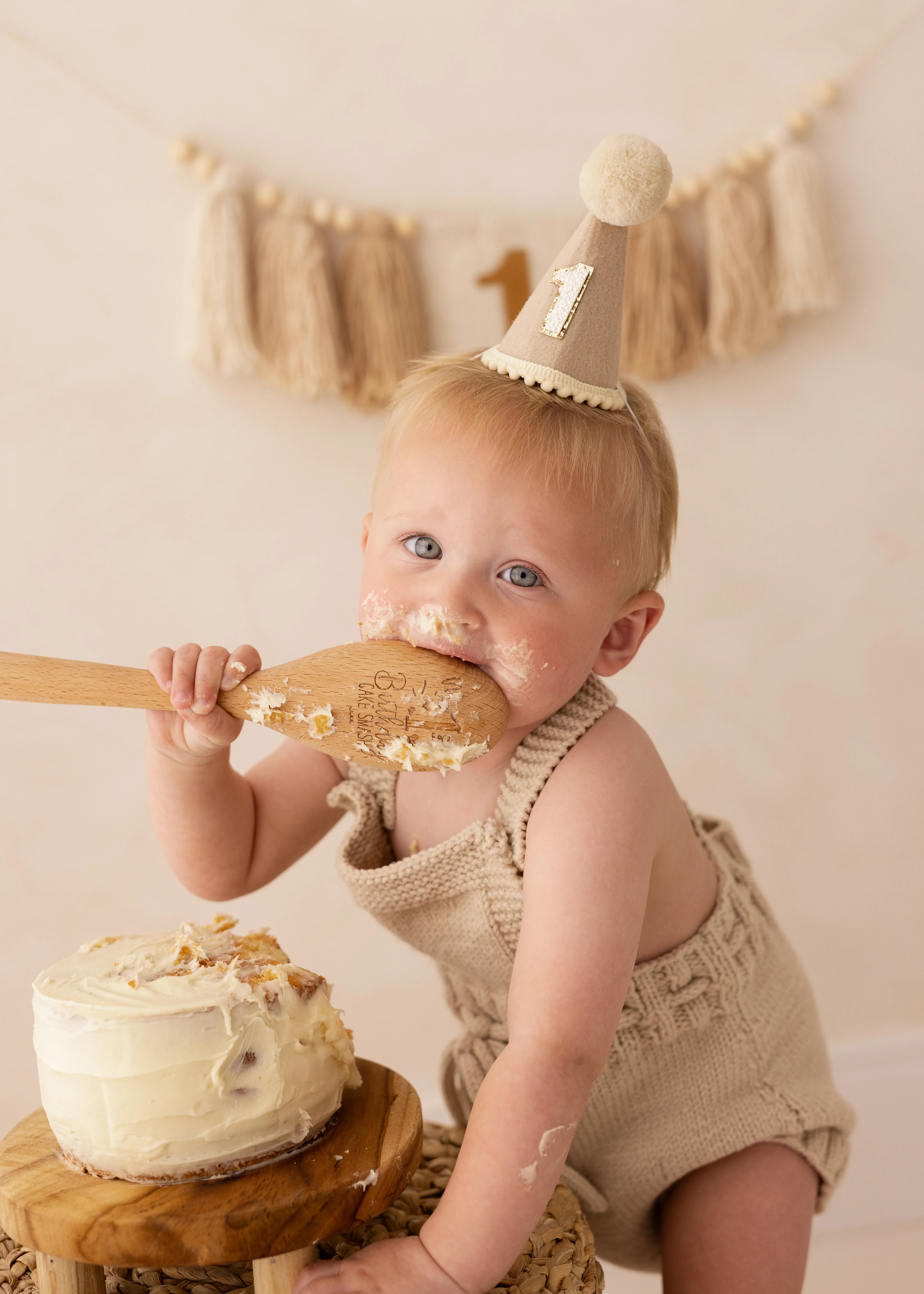 Baby wearing a “1” birthday hat and knitted romper, eating cake from a wooden spoon beside a small frosted cake during a fun cake smash photoshoot in Kent.