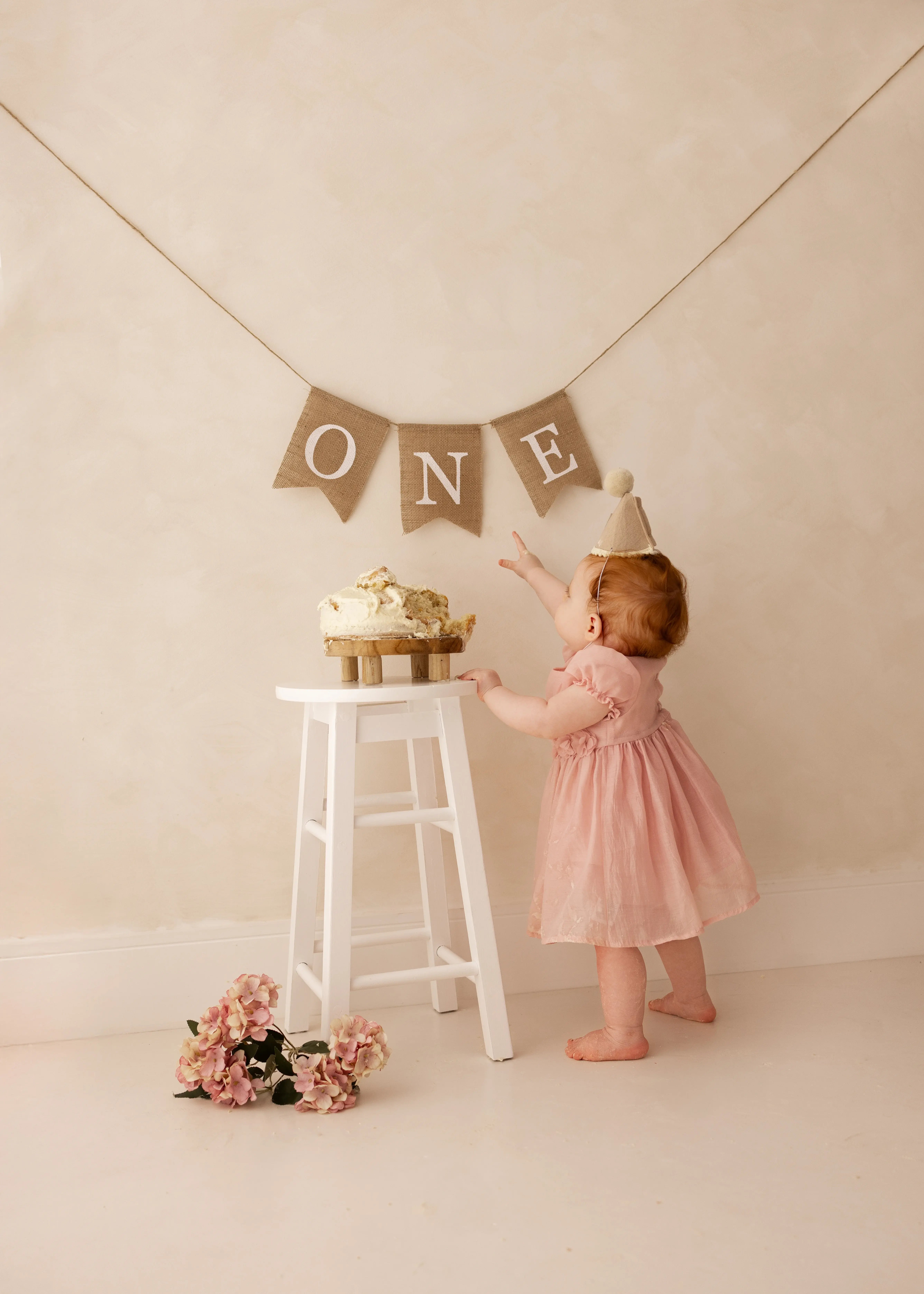 A toddler in a pink dress reaches up toward a “ONE” banner while standing beside a partly-smashed birthday cake on a white stool during a neutral, softly lit cake smash photoshoot.