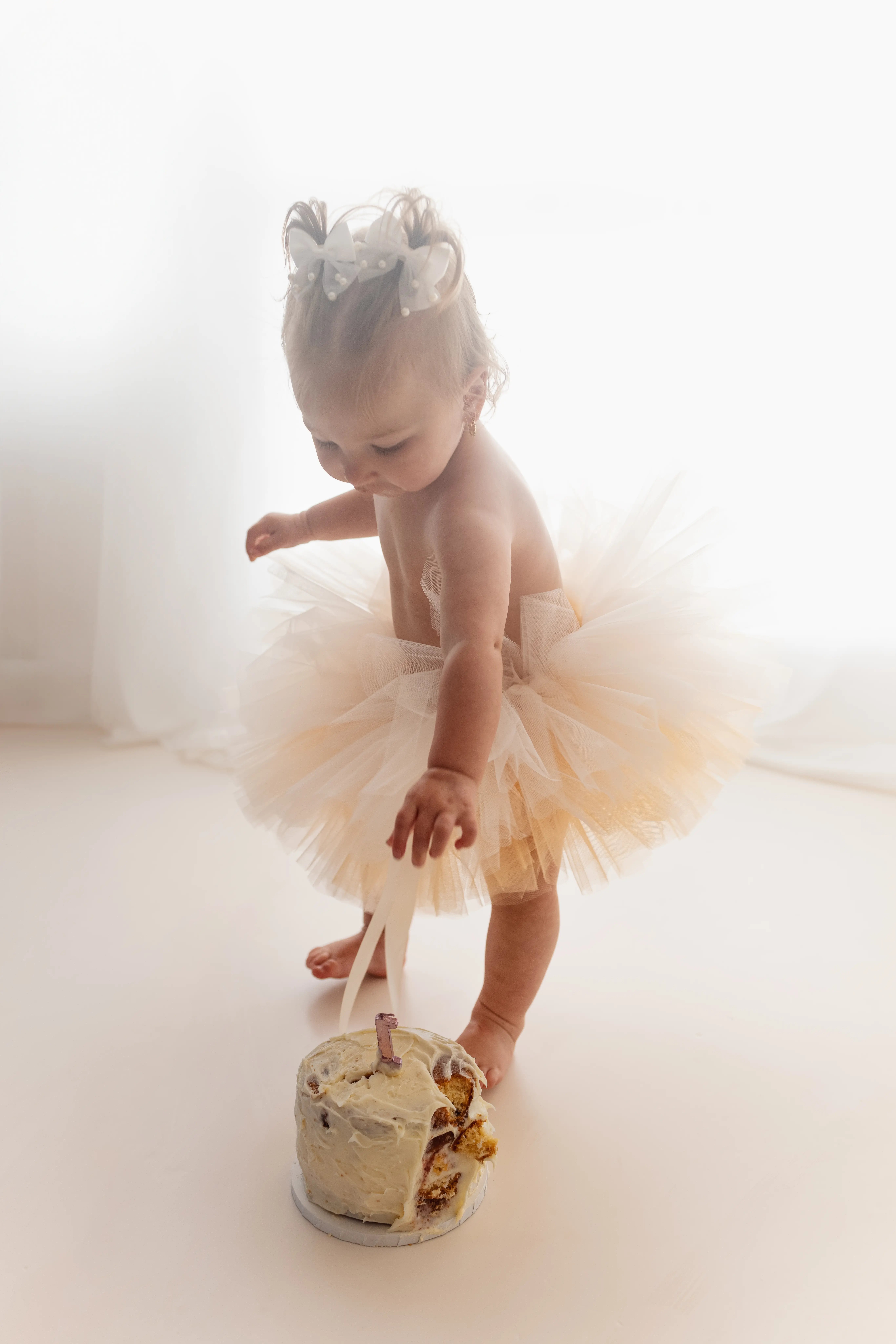 photograph of a 1 year cake smash, with the baby girl wearing a white tutu, standing over the cake
