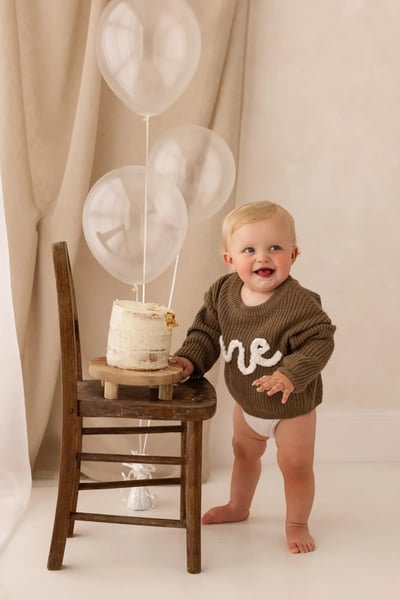 Smiling one-year-old standing beside a rustic chair with a first birthday cake and clear balloons, wearing a brown jumper that says 'one' during a cake smash photography session in Kent.