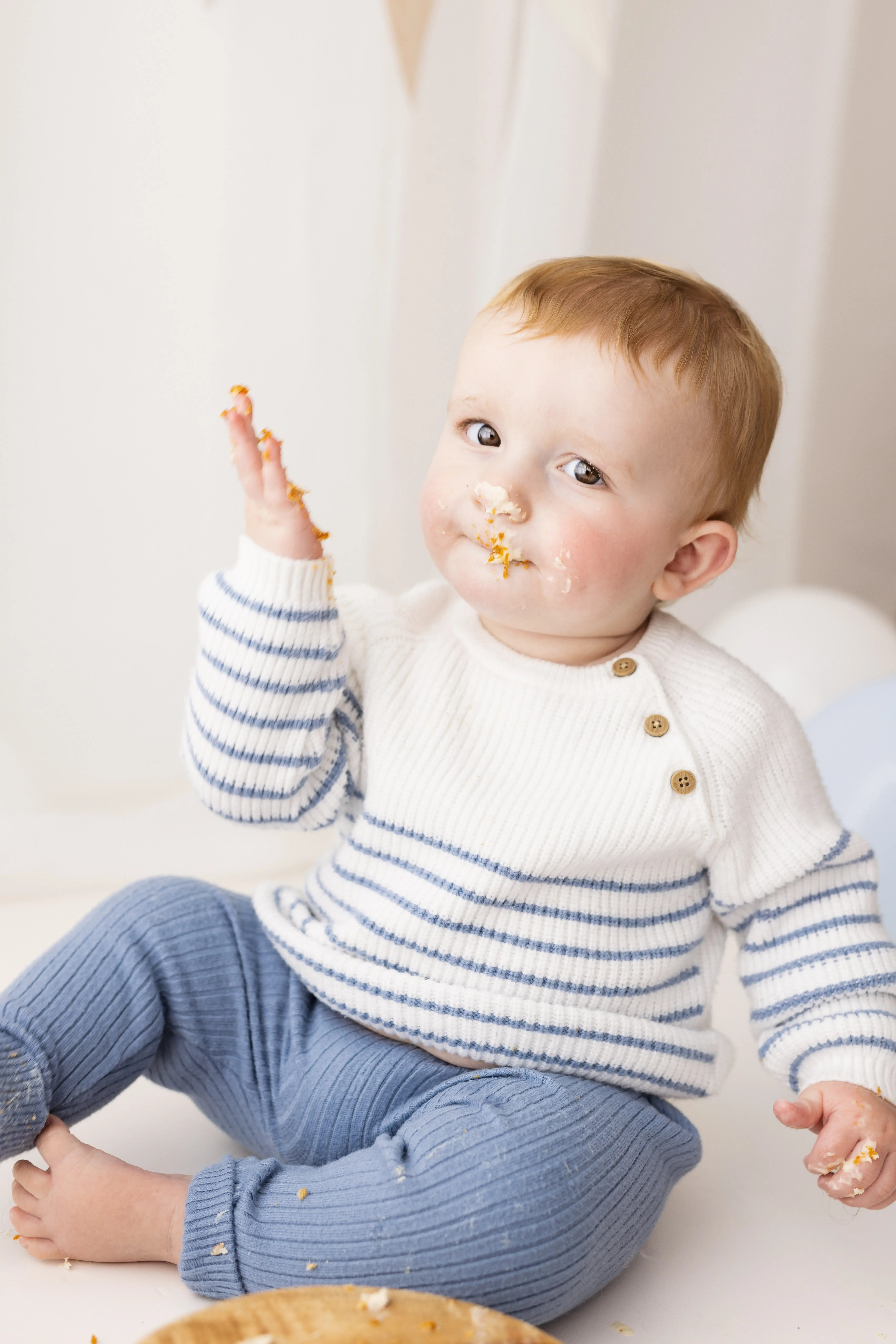 One-year-old boy dressed in a striped knit top, gleefully waving a frosting-covered hand during a playful first birthday cake smash, surrounded by white and pastel accents.