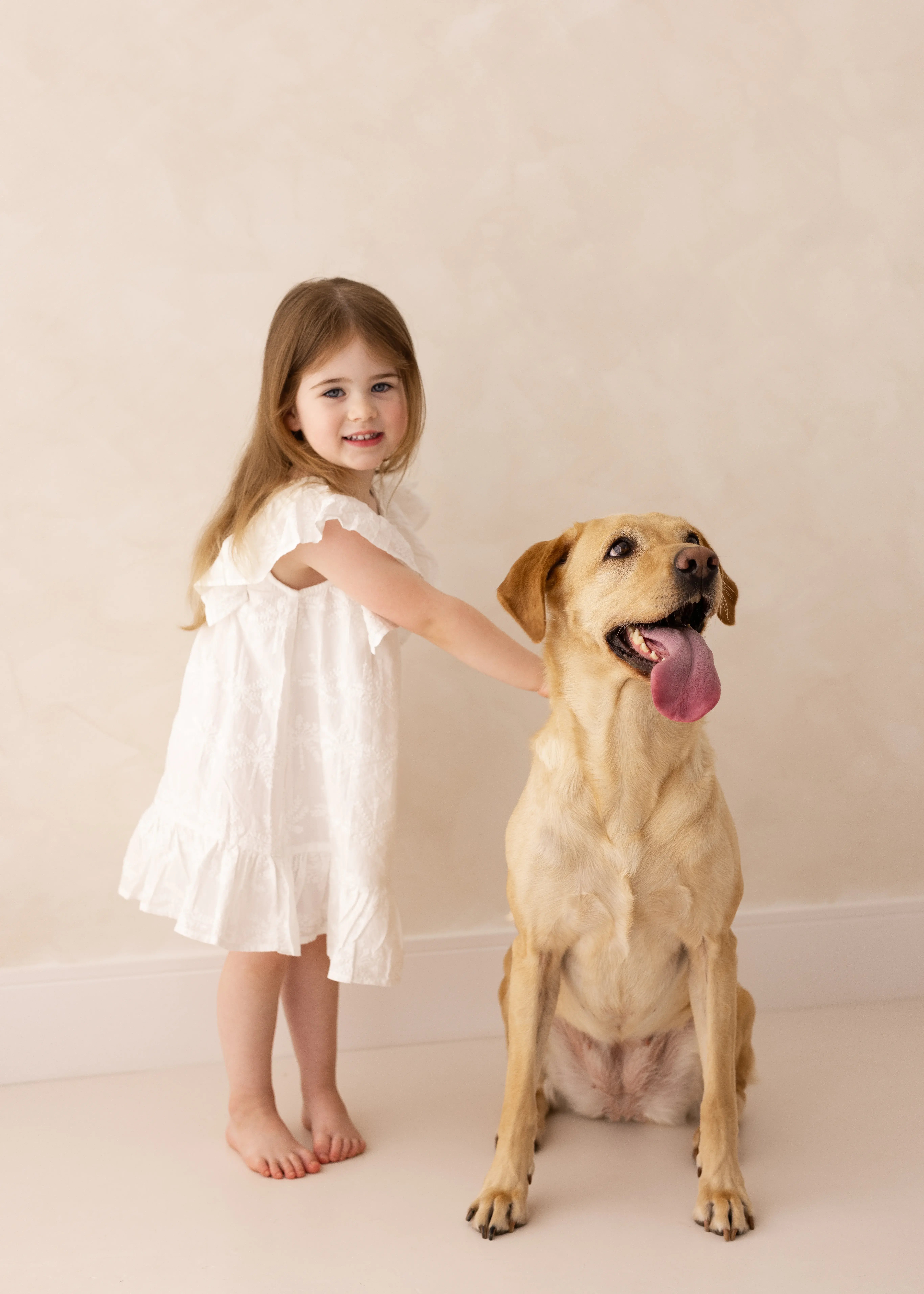 Young girl in a white dress smiling while gently holding a golden Labrador during a studio family photography session.