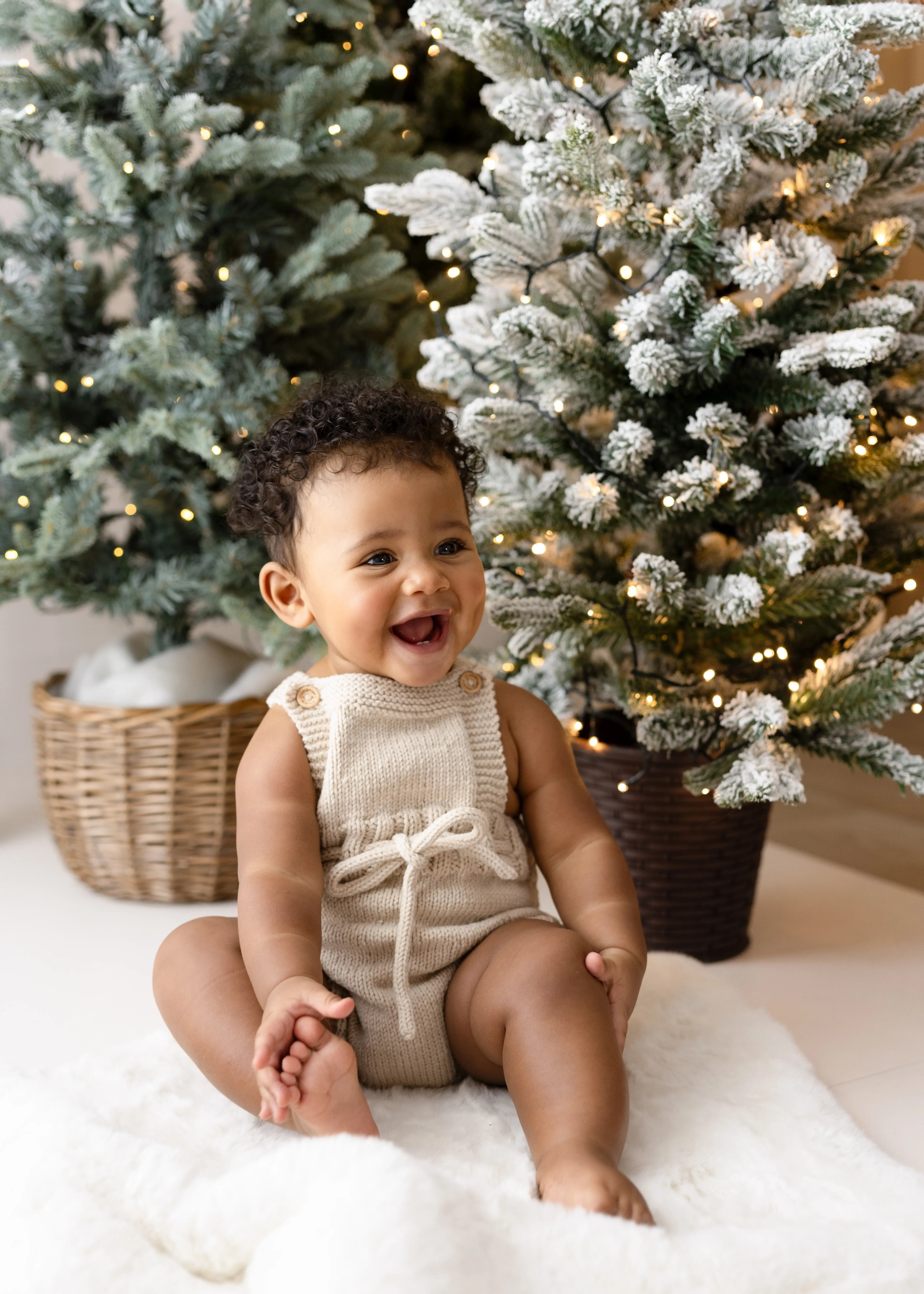 Delighted baby boy wearing a cosy beige romper, smiling widely among snow-frosted Christmas trees adorned with fairy lights in a warm holiday mini shoot.