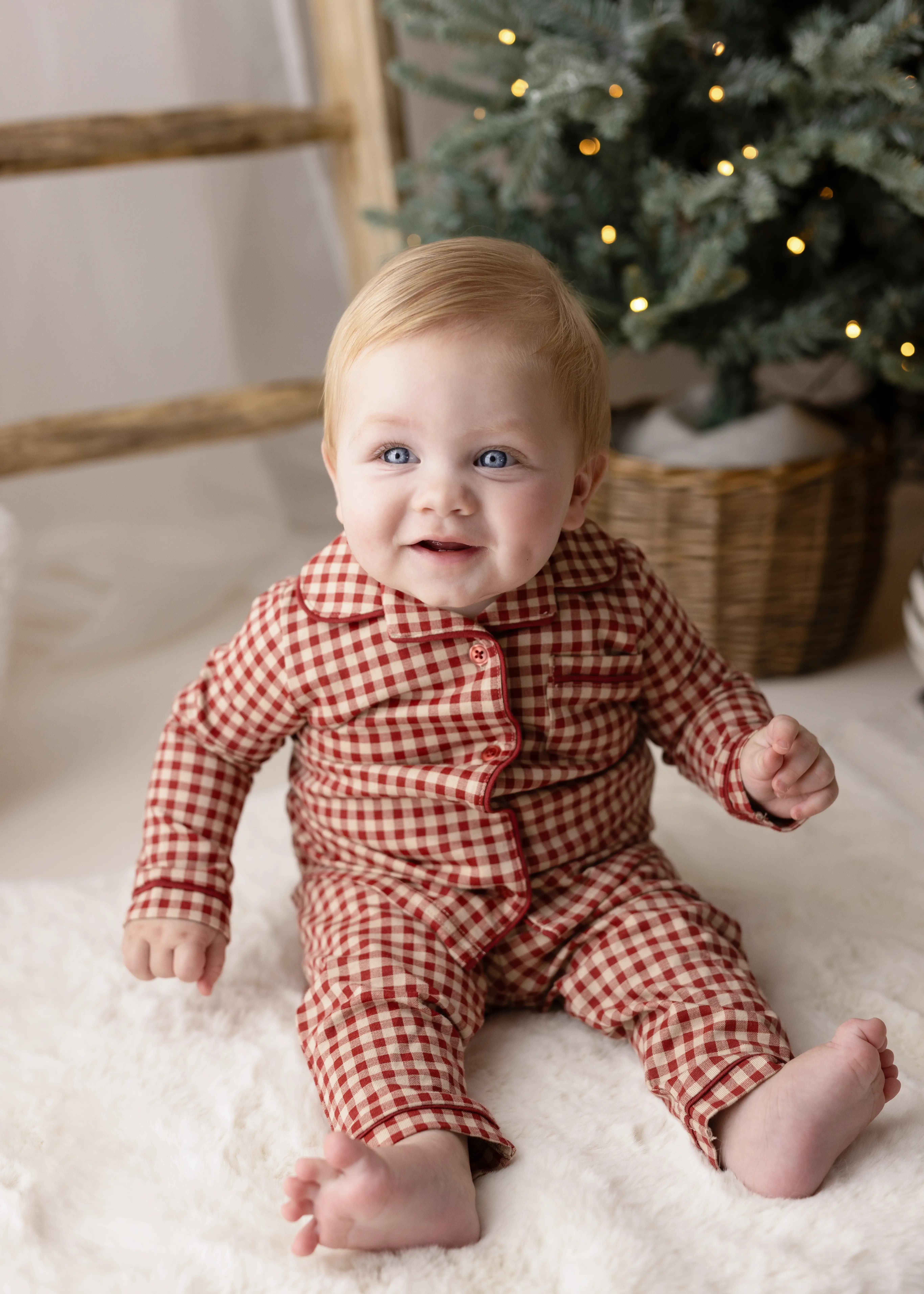 Smiling baby boy in cosy red gingham pyjamas, seated on a fluffy white rug by a softly lit Christmas tree, capturing a warm and festive holiday mini portrait.