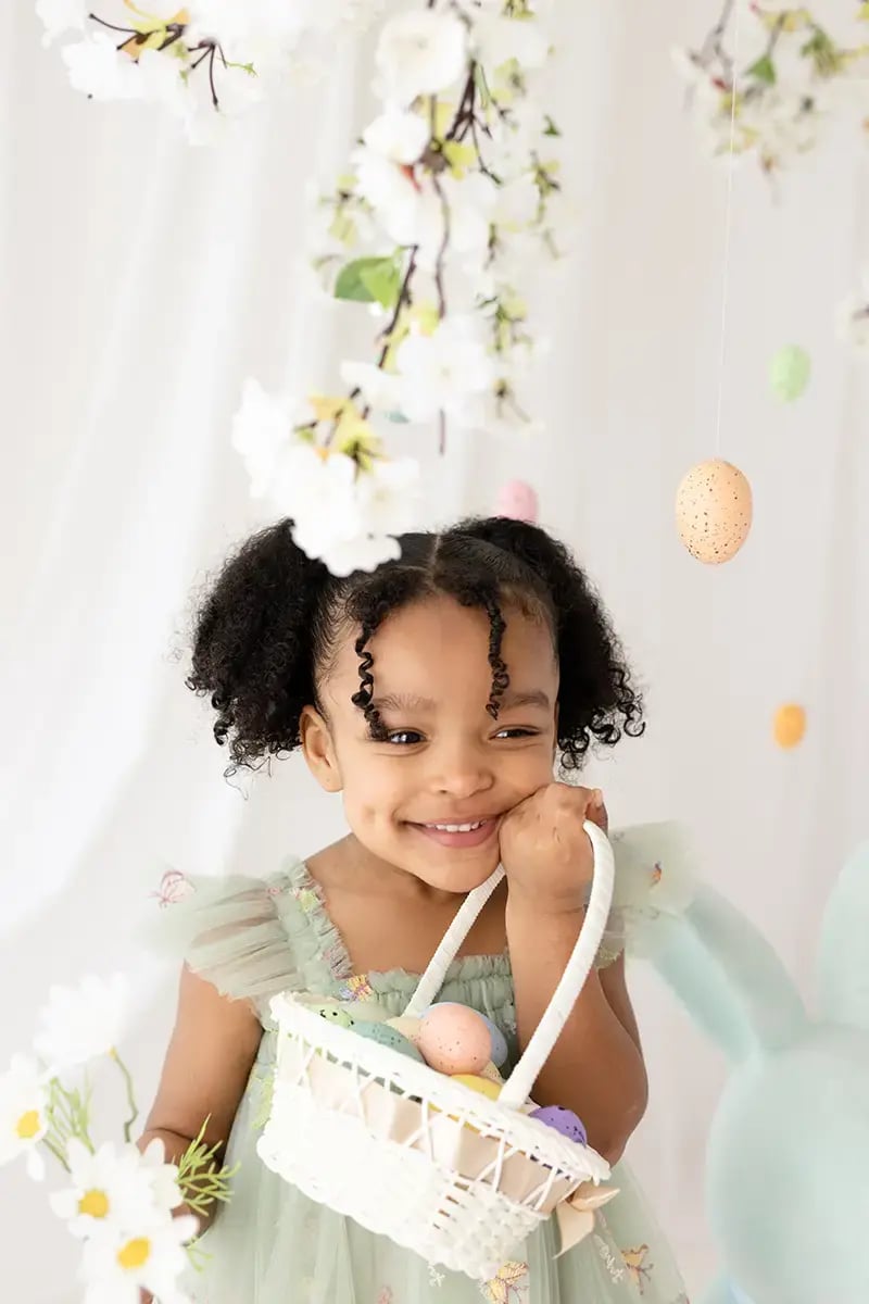 Close-up of a smiling little girl in a pale green dress holding a white Easter basket of painted eggs, surrounded by hanging blossoms in a spring children’s photoshoot.