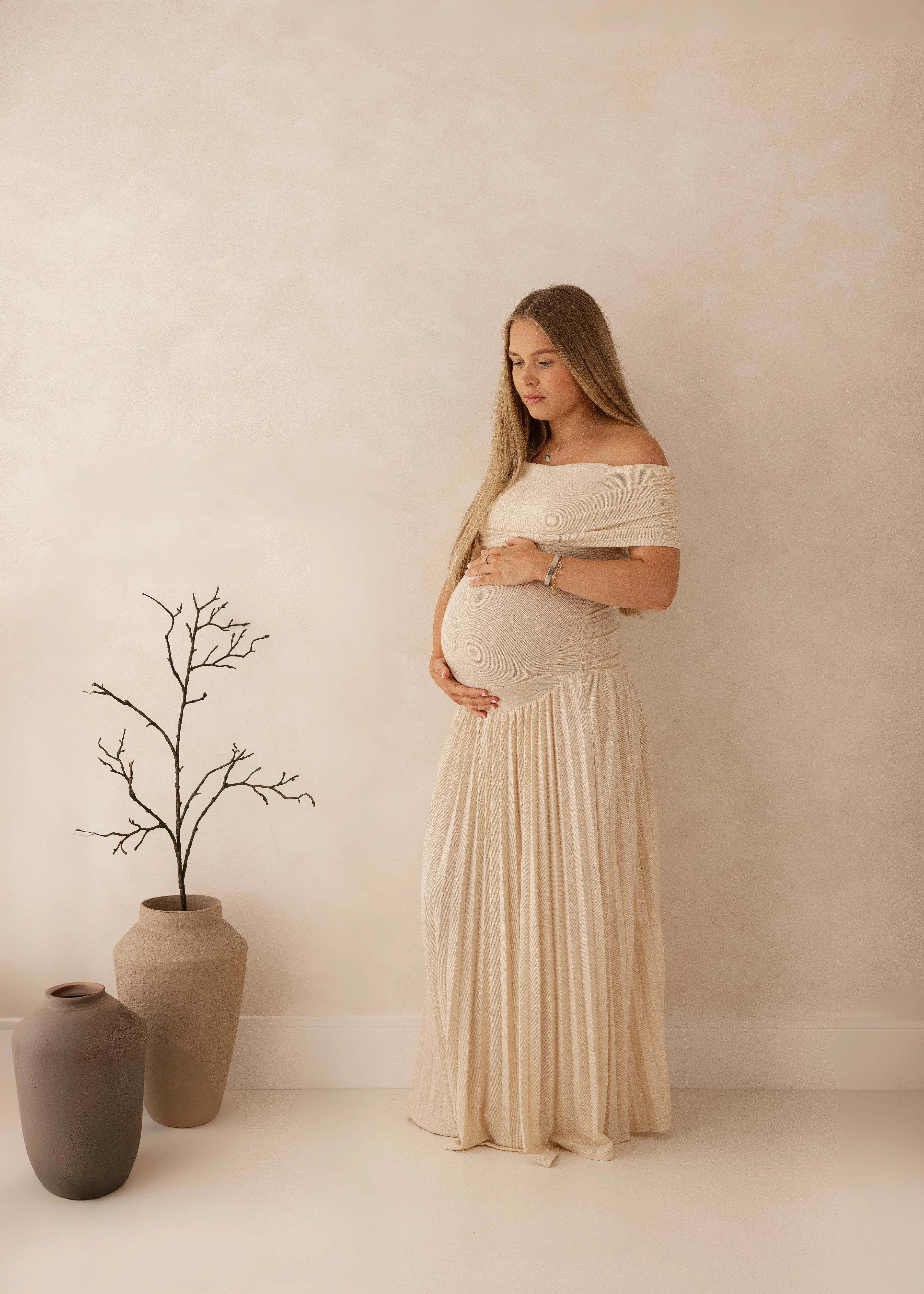 Pregnant woman in a flowing neutral gown holding her baby bump during a serene maternity photoshoot in a soft, minimal studio setting in Kent.