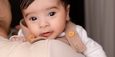 Photograph of a baby ready for their newborn photoshoot, over their parents' shoulder looking into the camera.