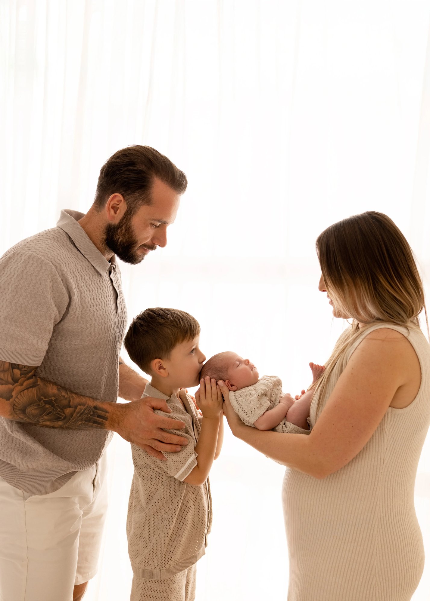 Parents and older brother leaning in as the brother kisses his newborn sibling’s head while the baby rests in their mother’s arms during a soft, natural family newborn photoshoot in Kent.