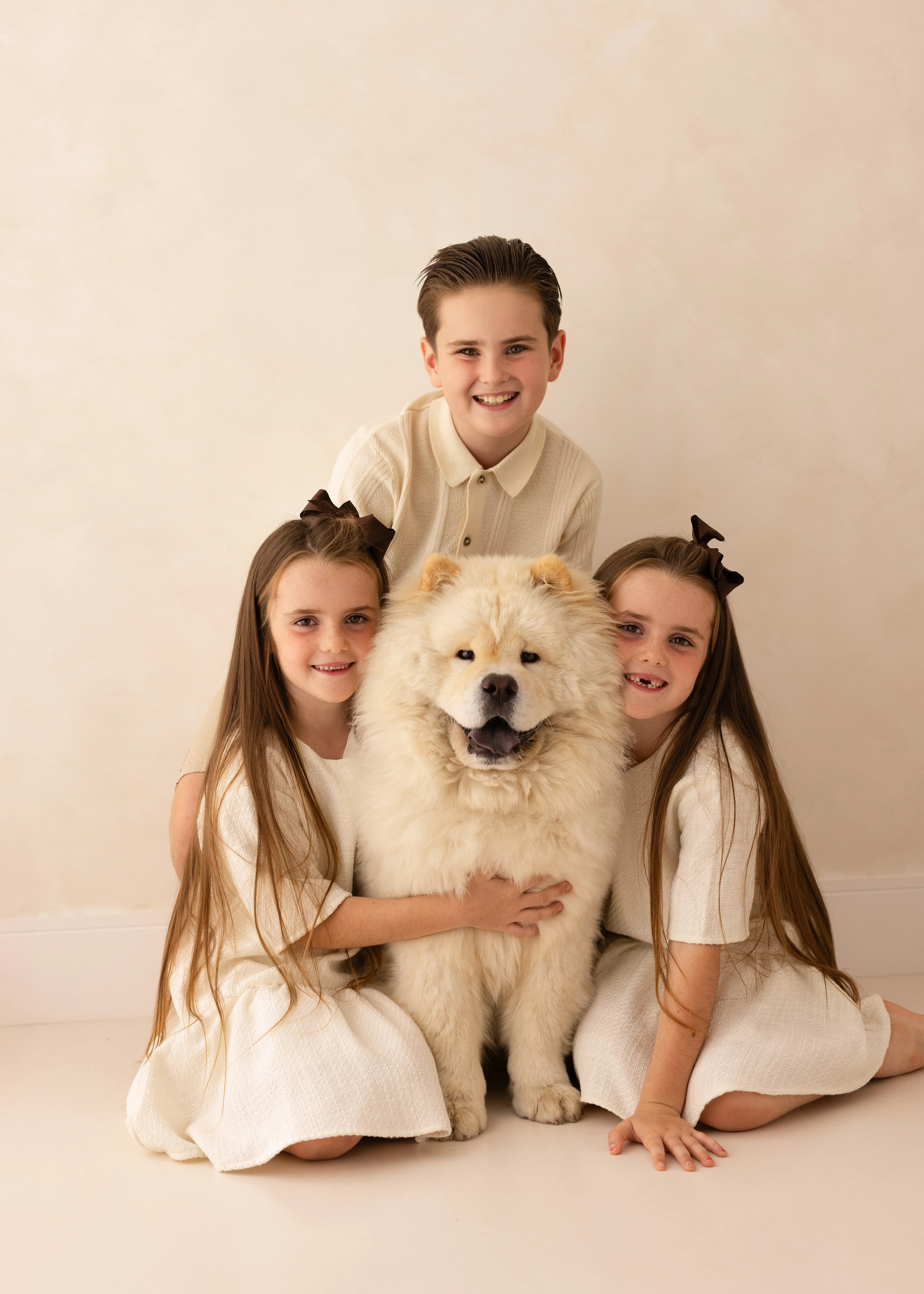Three smiling children sit closely around their fluffy cream Chow Chow dog during a studio family portrait. The boy kneels behind while the two girls in matching cream dresses cuddle the dog from either side, all framed against a soft beige backdrop.