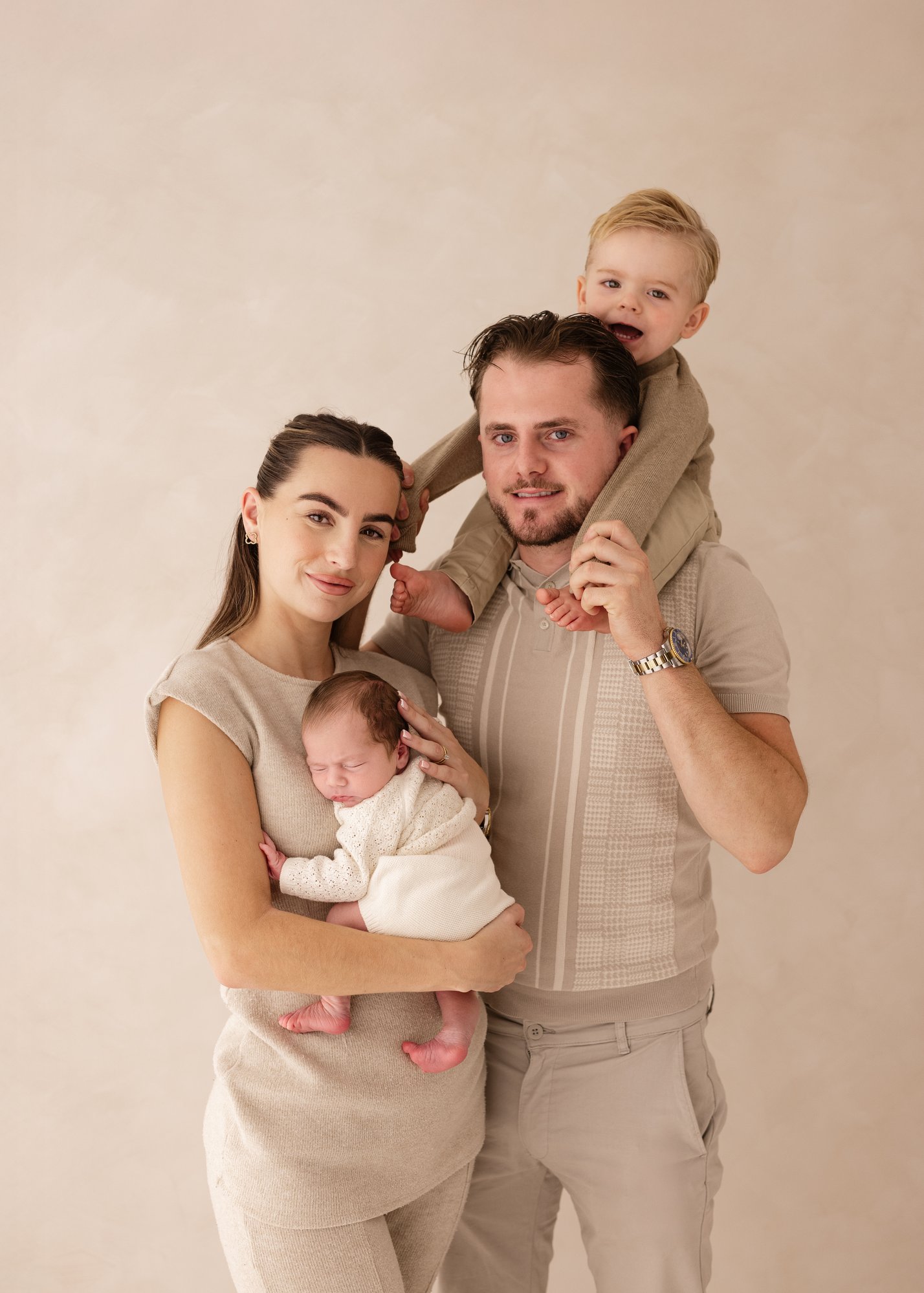 Parents holding their newborn baby while their toddler sits on the father’s shoulders during a soft, neutral-toned family newborn photography session in Kent.