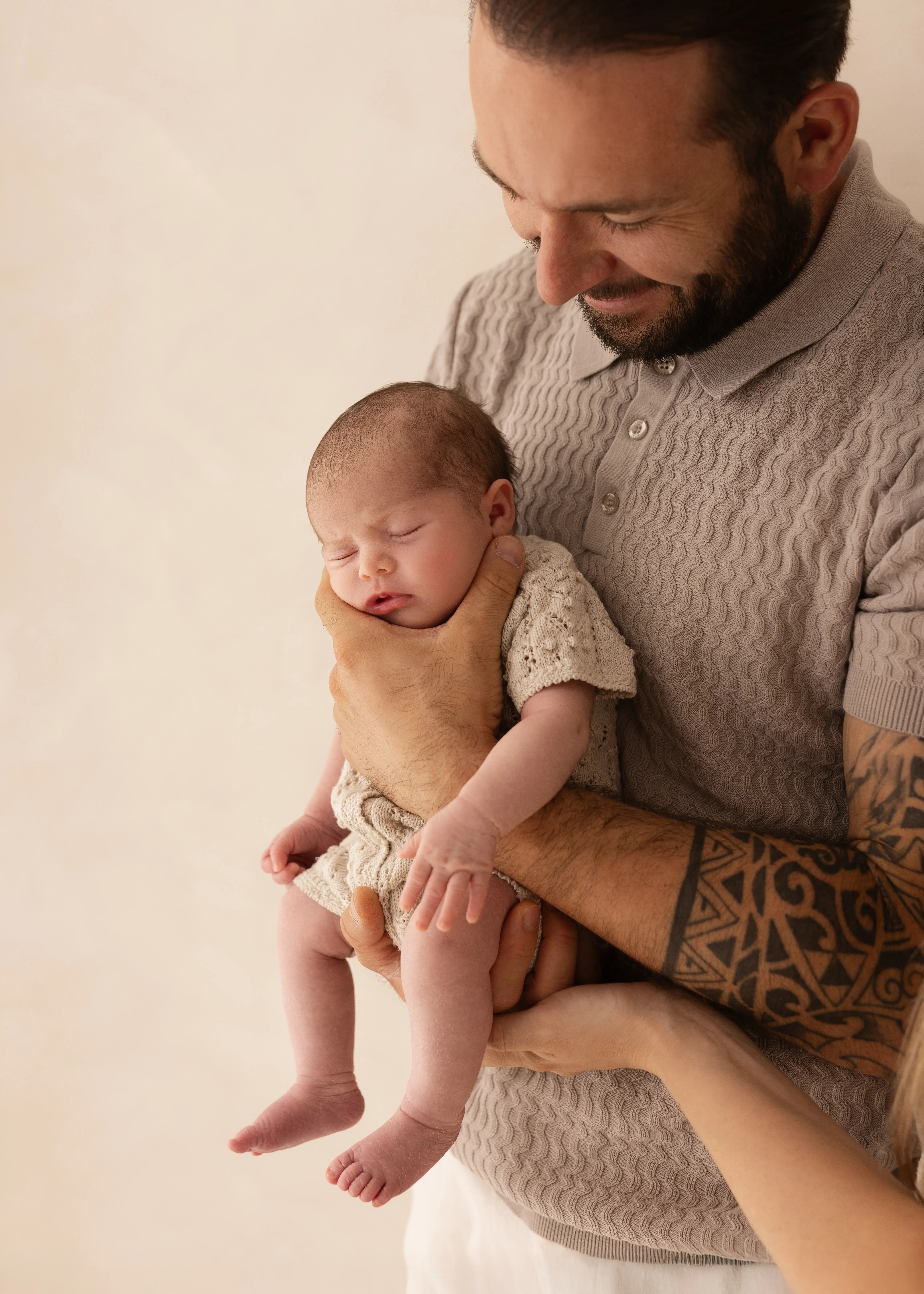 Father smiling down at his sleeping newborn baby, holding them gently in a cream knitted outfit during a natural newborn photoshoot in Kent.