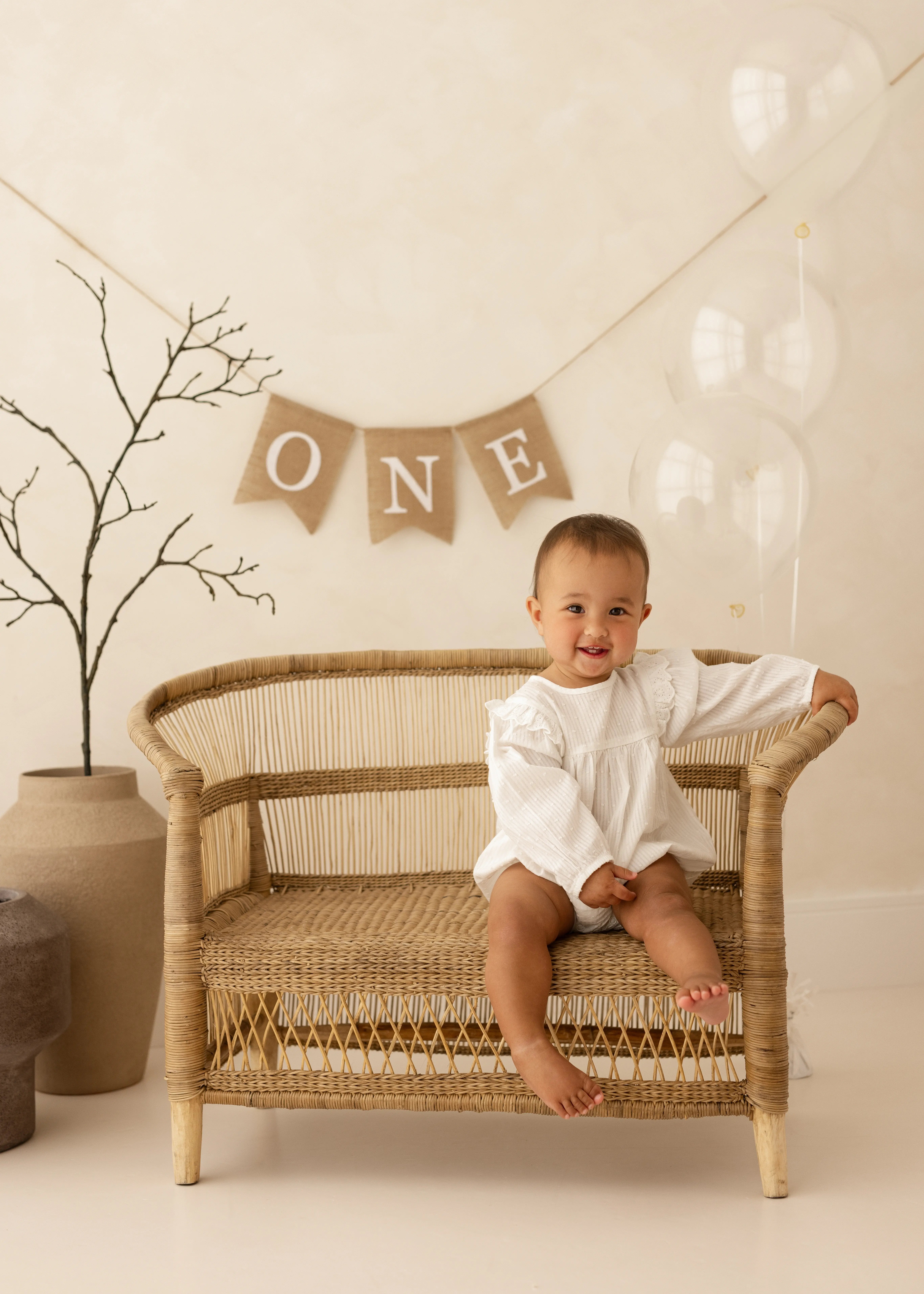 One-year-old girl sitting on a wicker bench during her first birthday photoshoot, with a neutral “ONE” banner and clear balloons in the background.