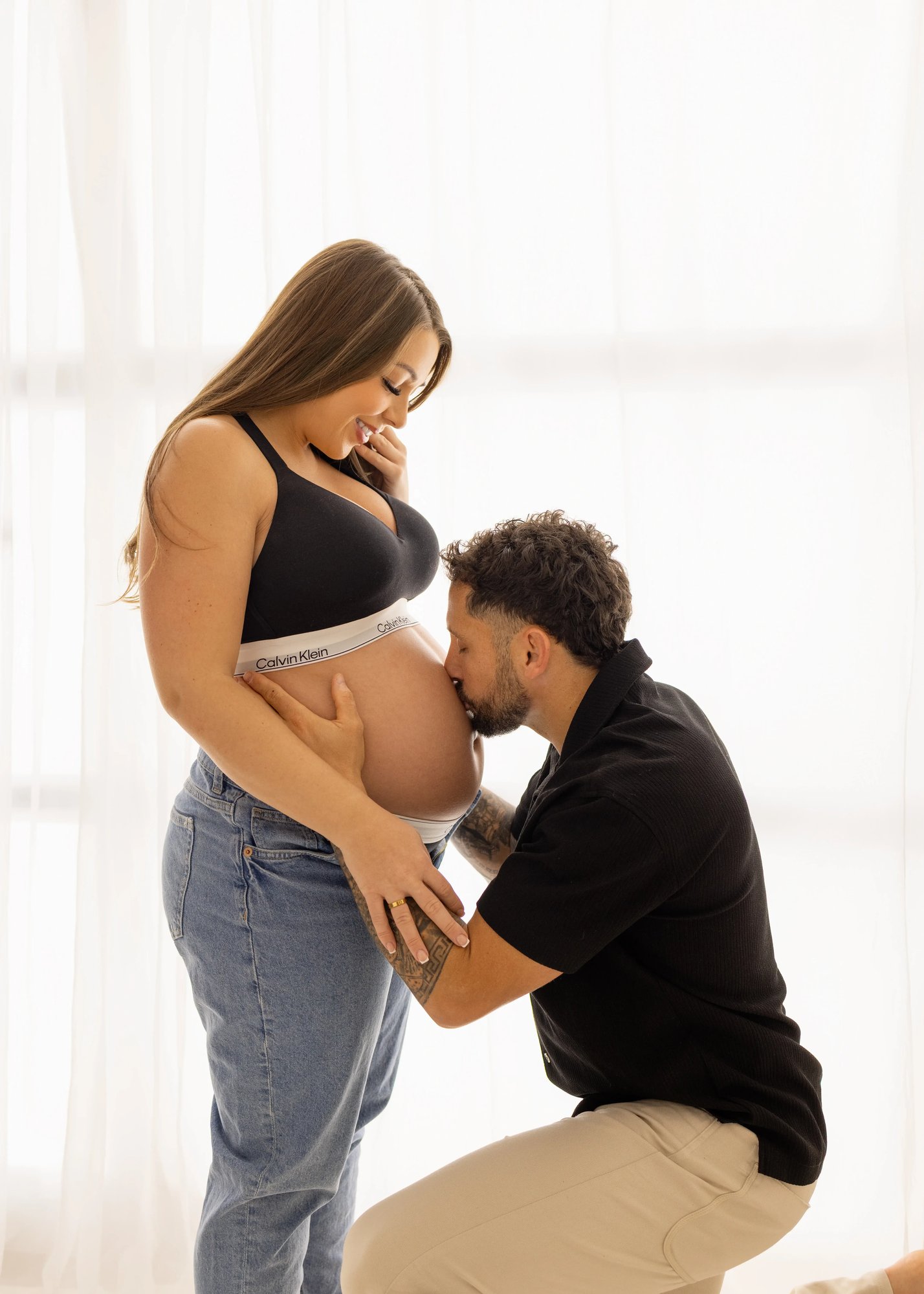 Expectant couple during a maternity photoshoot, with the father gently kissing the mother’s baby bump as she smiles, captured in soft natural studio light in Kent.