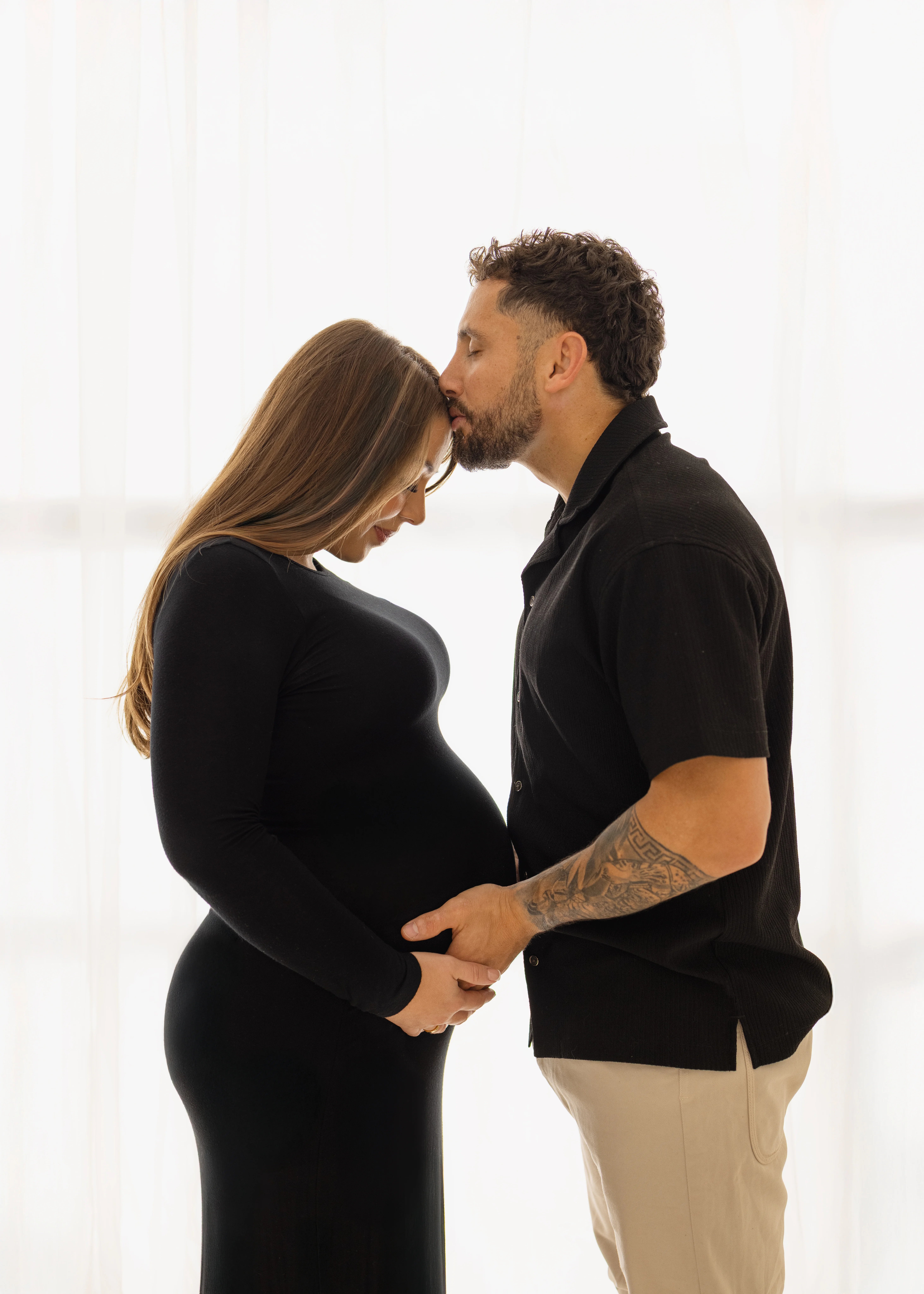 Expectant couple standing close together as the partner kisses her forehead and both rest their hands on the baby bump, captured in soft backlit studio light during a maternity photoshoot in Kent.