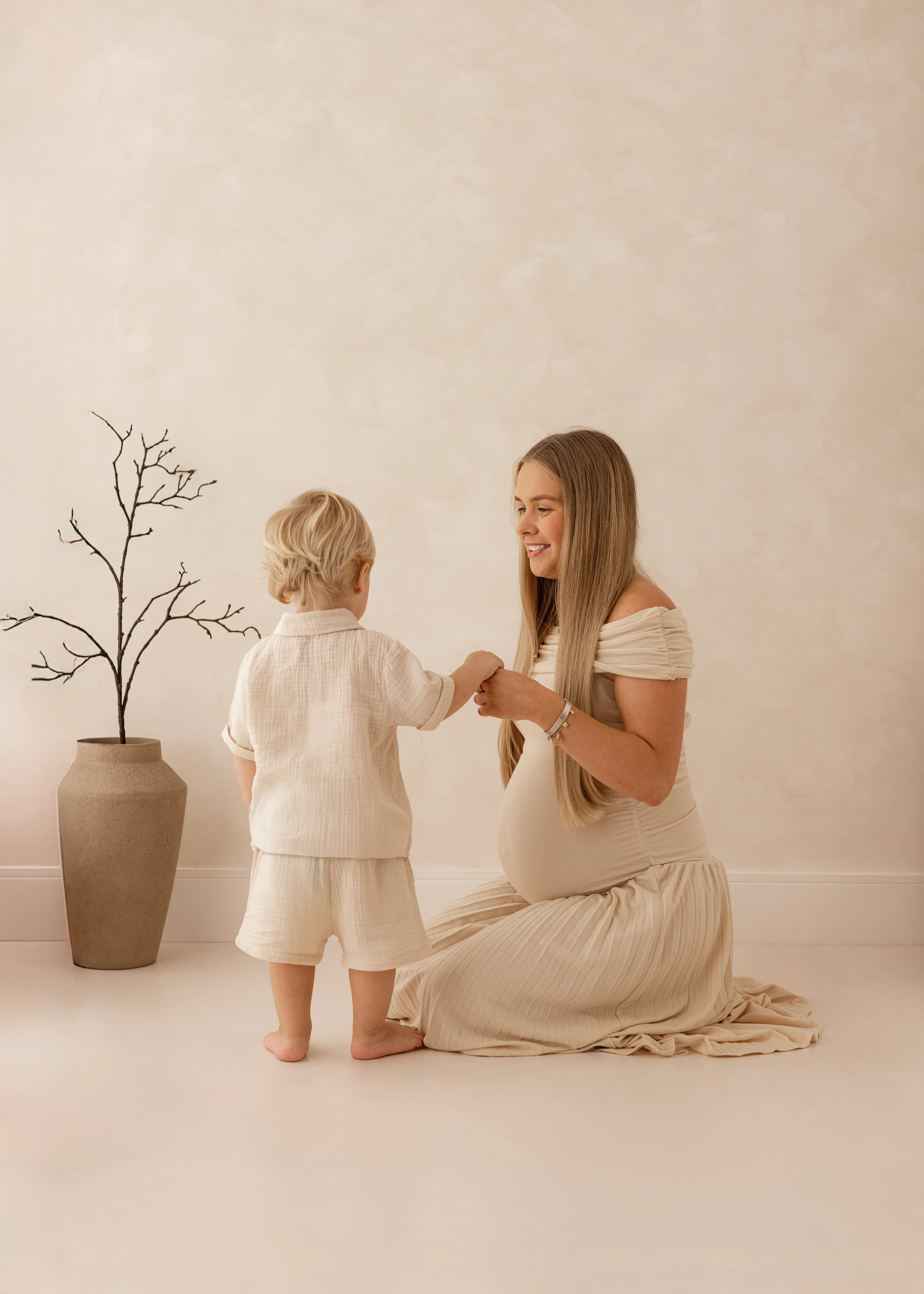 Mother holding hands with her young toddler during a maternity photoshoot, both dressed in soft neutral outfits, standing beside a minimalist vase and branch decoration in a natural light studio.