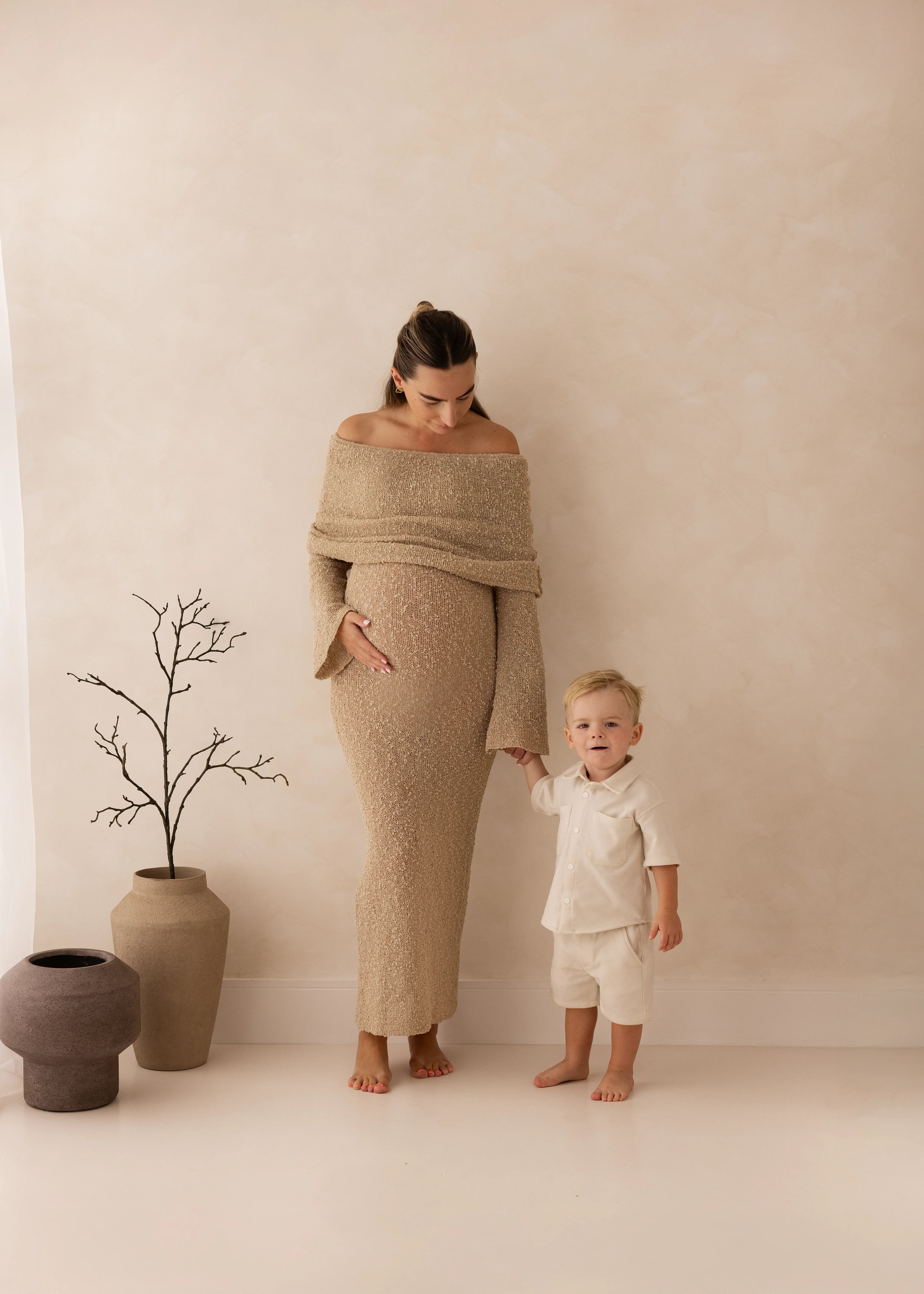 Pregnant mother in a textured neutral gown holding her toddler’s hand during a soft, minimalist maternity photoshoot in Kent.
