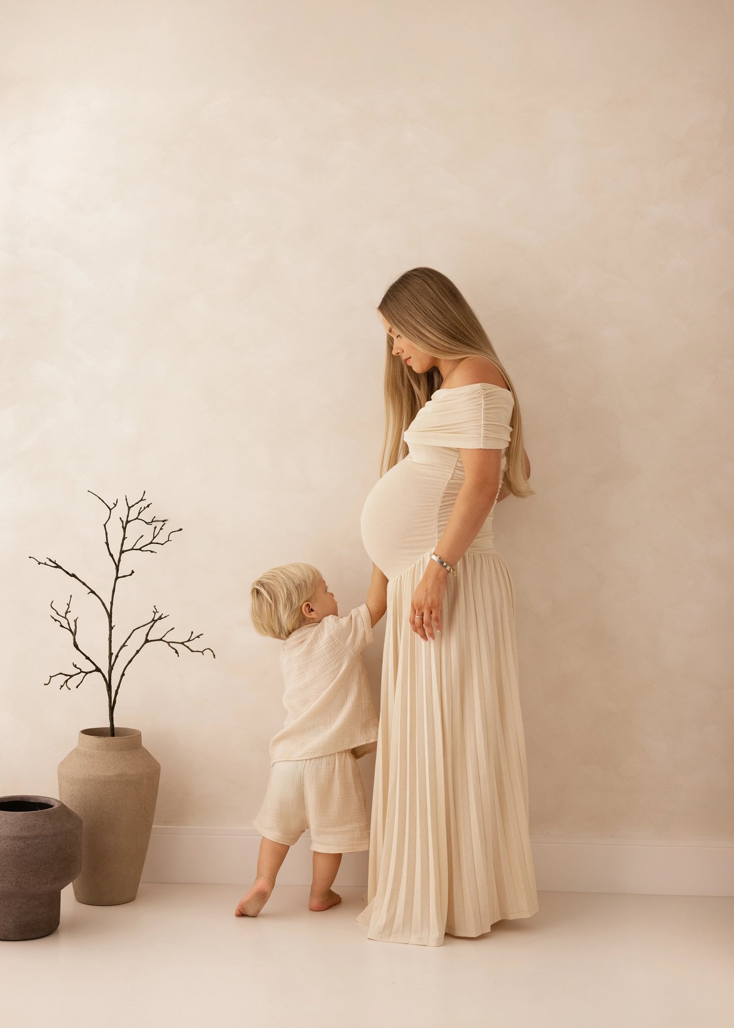 Pregnant mother in a neutral off-shoulder dress holding her toddler’s hand as he reaches up toward her baby bump during a soft, natural maternity photoshoot in Kent.