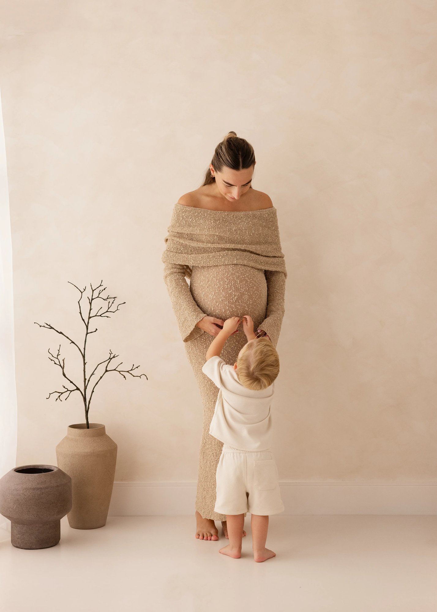 Toddler reaching up to touch his pregnant mother’s baby bump while she looks down lovingly, captured in a soft neutral studio during a maternity photoshoot in Kent.