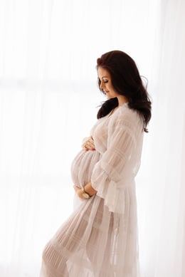 Graceful expectant mother in a sheer white lace gown, standing in a sunlit studio and tenderly holding her bump for an elegant, airy maternity photo session.