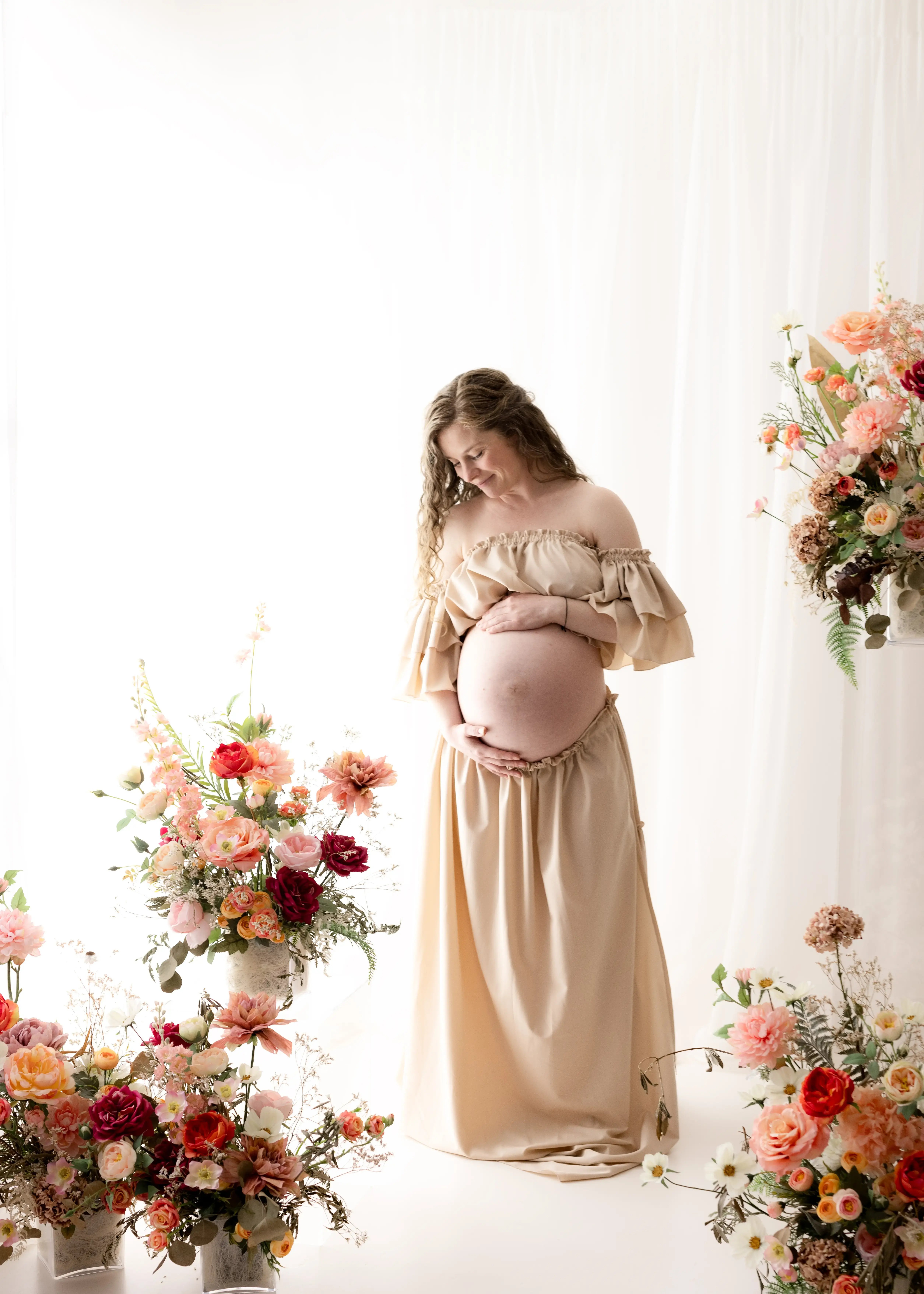 Side-view maternity portrait of a smiling woman in a ruffled beige dress, standing among vibrant spring flowers and basking in natural light for a dreamy floral maternity photography session.