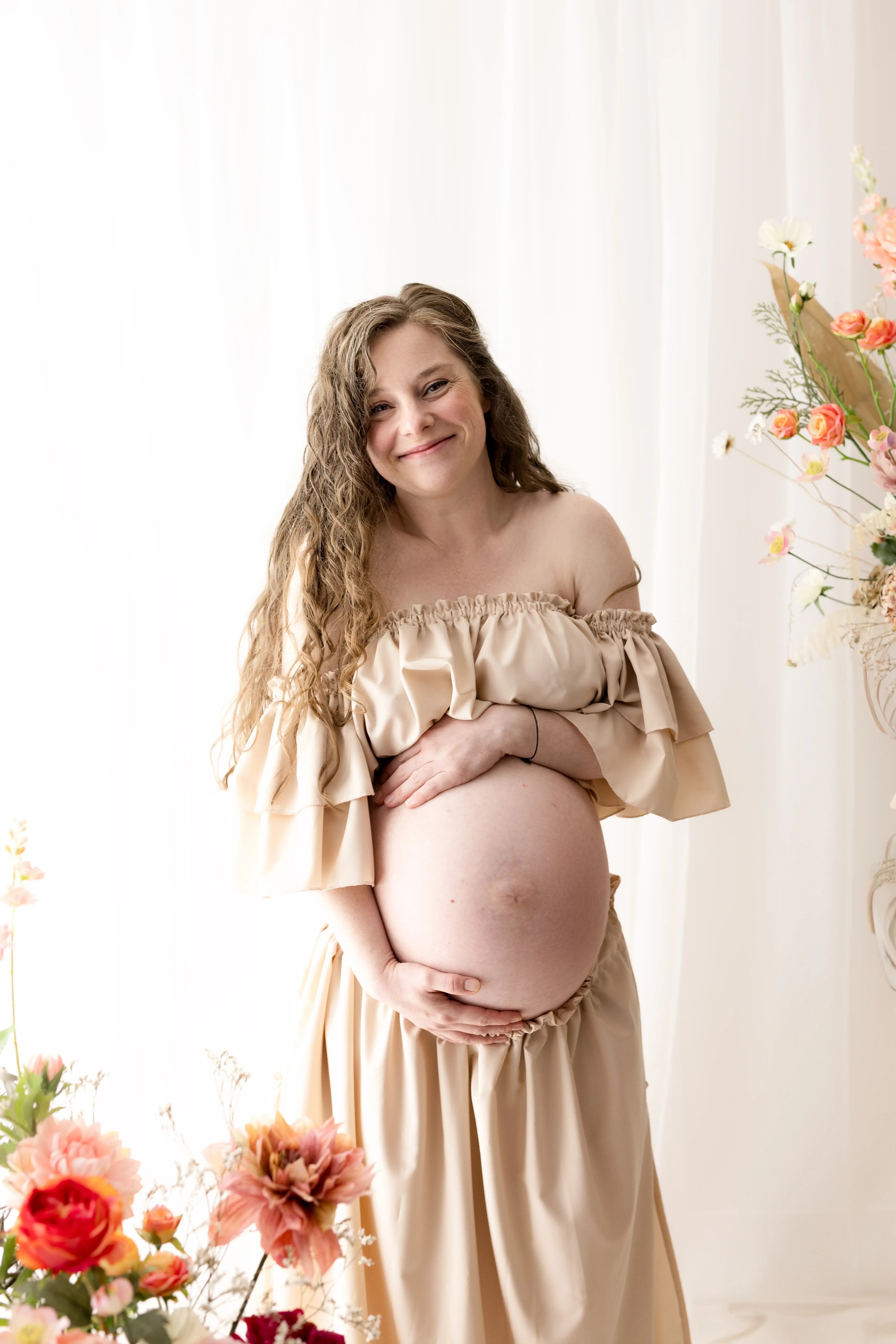 Radiant expectant mother in a flowy, off-the-shoulder beige gown gently cradling her baby bump amid soft pastel blooms, capturing a warm and intimate moment of floral maternity photography.