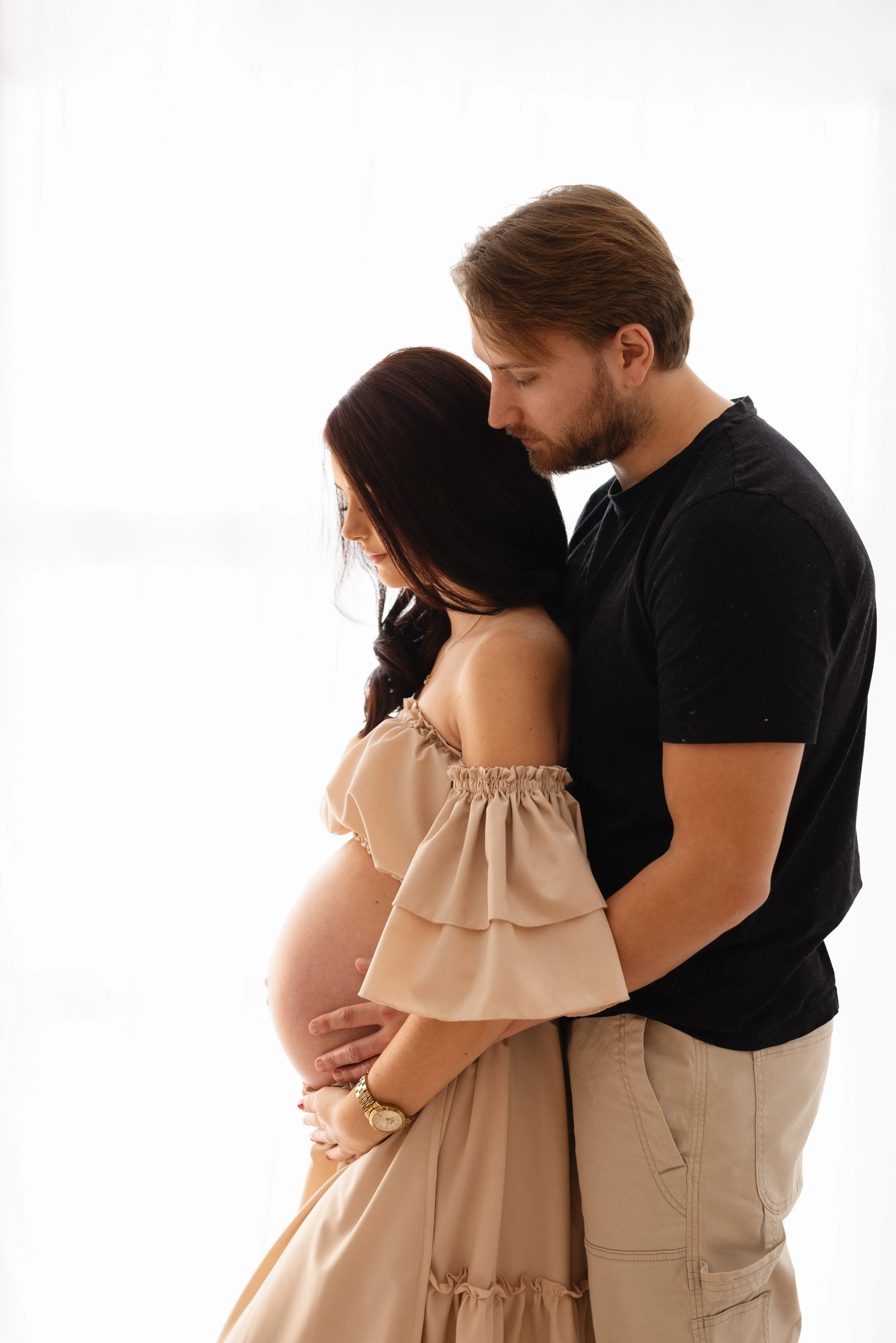 Couple’s intimate maternity portrait featuring a mother-to-be in an off-the-shoulder beige gown, gently embraced from behind by her partner in a softly lit, minimalist studio setting.