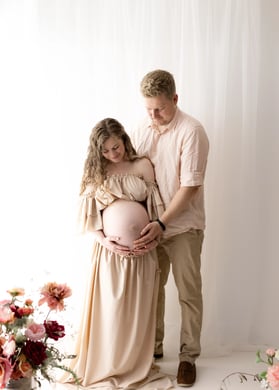 Tender moment between a pregnant woman in a flowing beige gown and her partner, both softly smiling and holding her baby bump while surrounded by romantic floral arrangements during their maternity photography session.