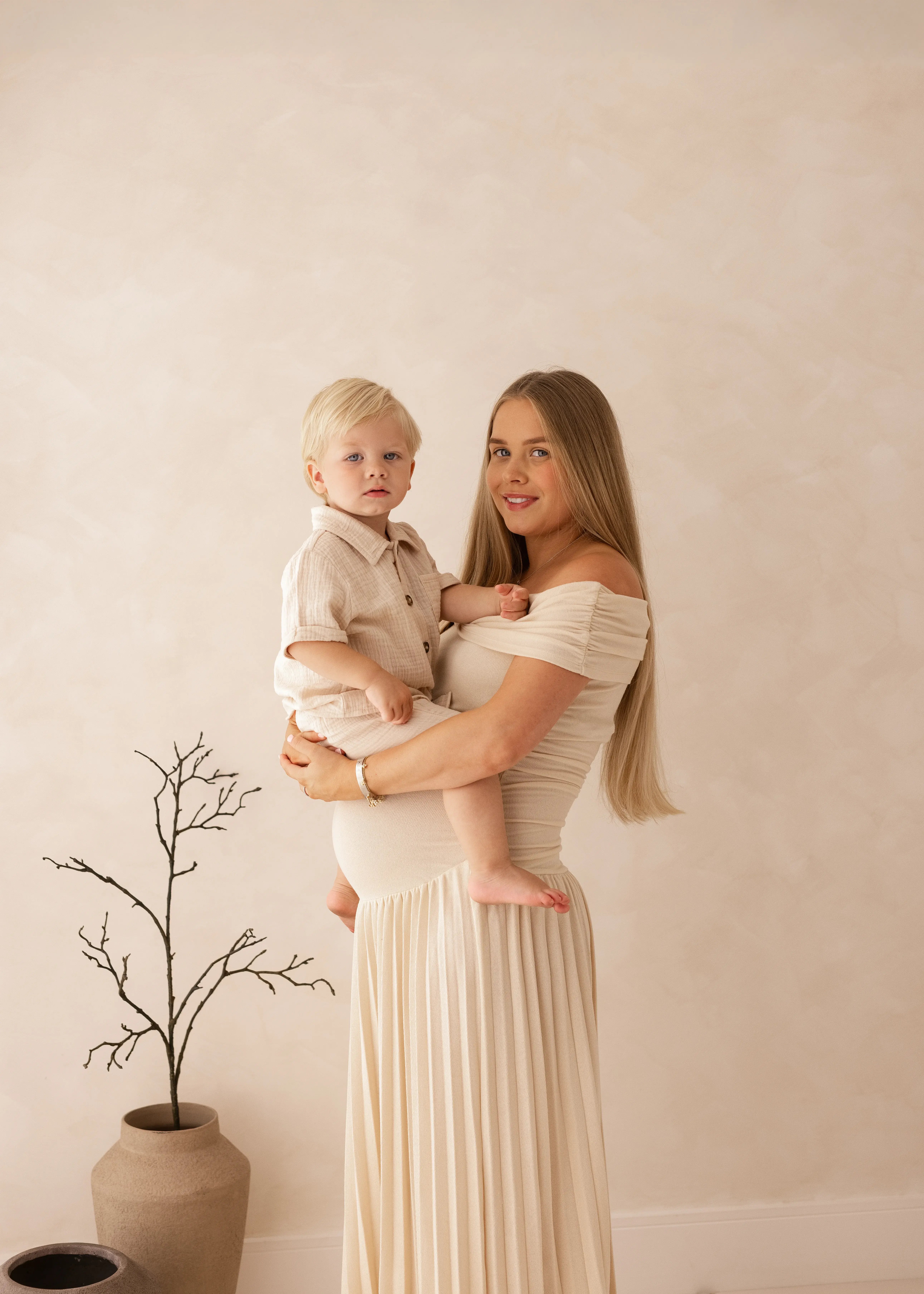 Mother holding her toddler while standing in a soft, neutral studio setting, both dressed in light tones, during a natural mother and child photography session in Kent.