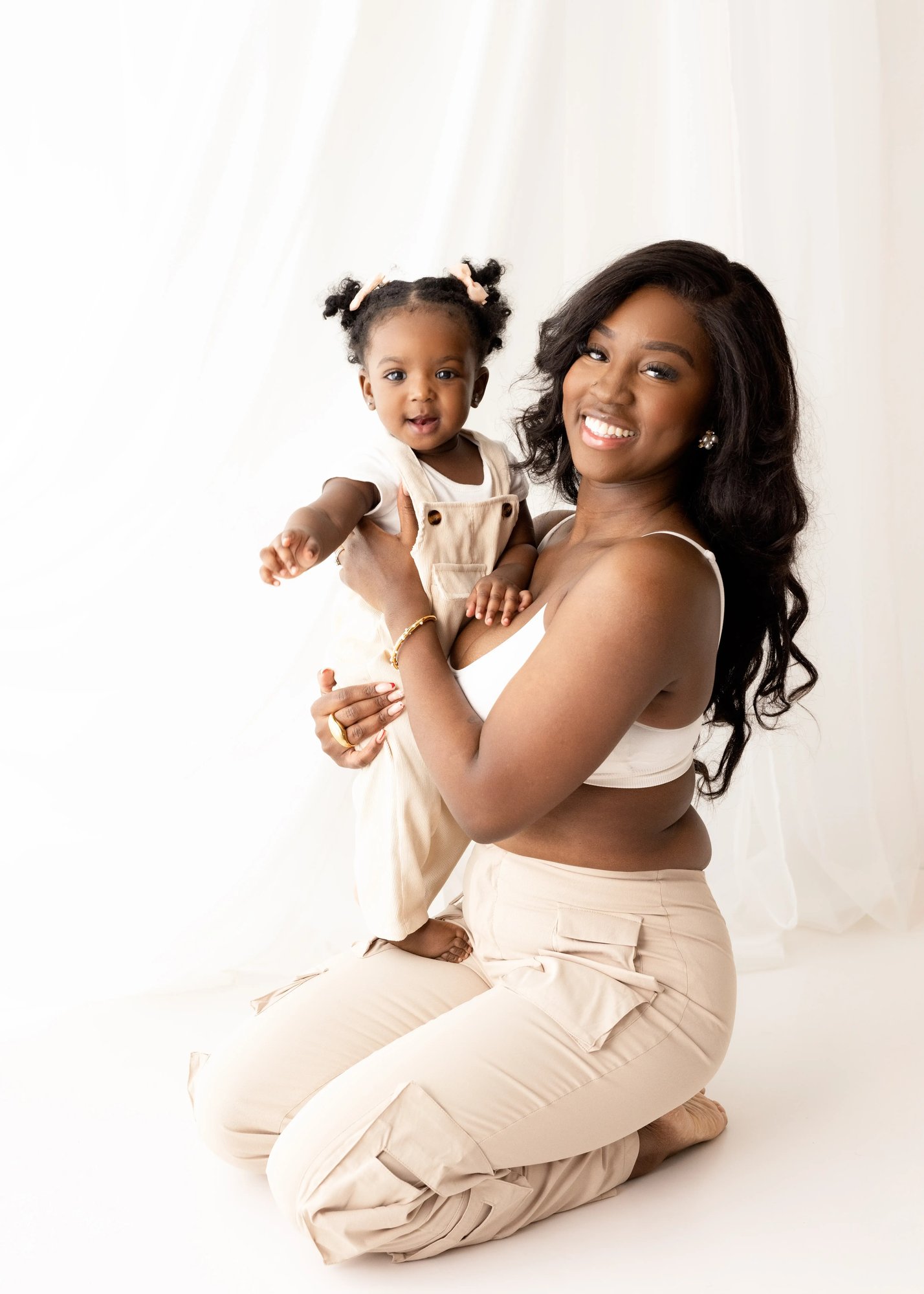 Mother in a white crop top and neutral trousers holds her toddler daughter dressed in beige overalls, both smiling radiantly in a softly lit, minimalist studio for a mummy and me mini session.