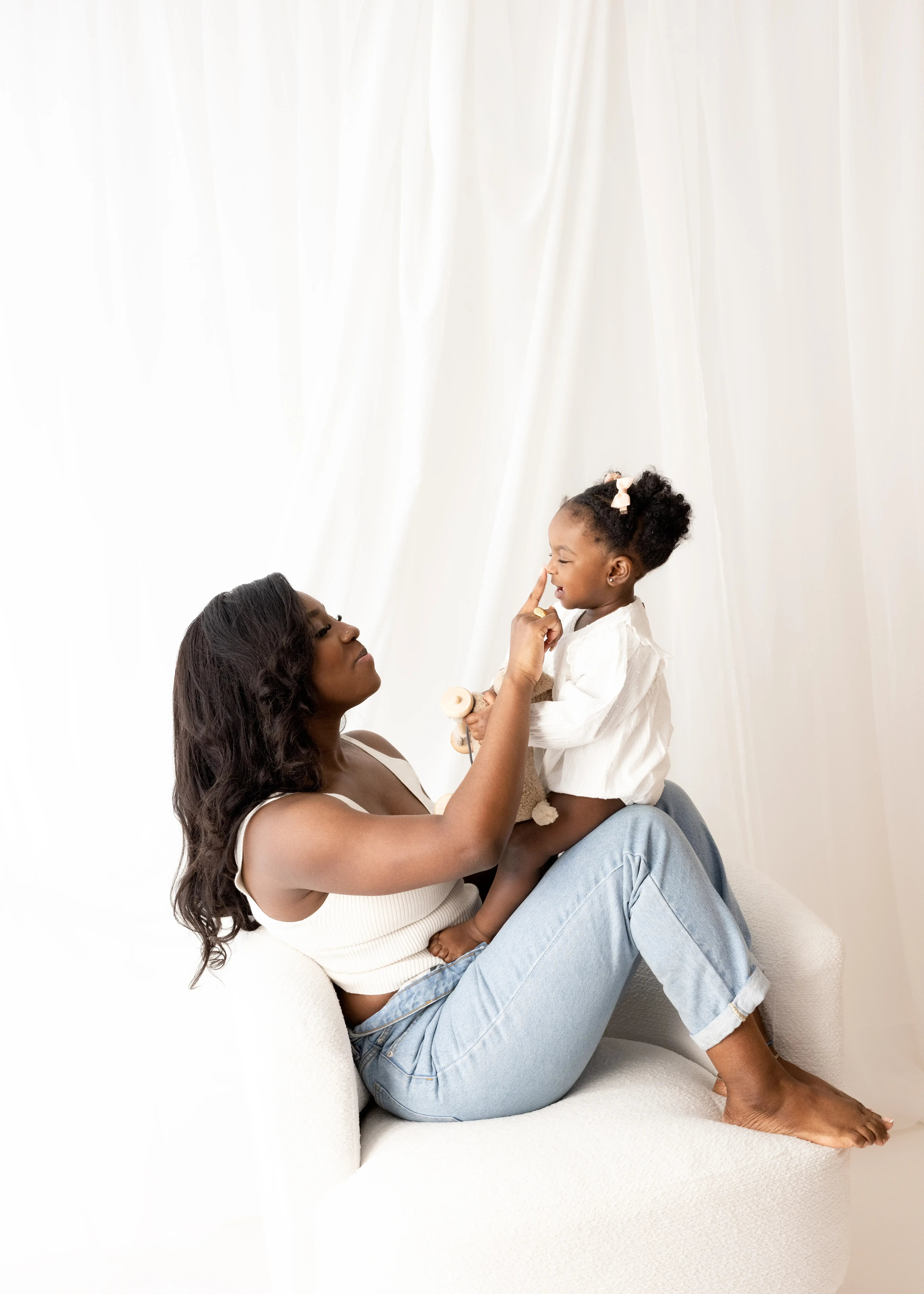 Mum relaxing on a soft chair, playfully tapping her toddler daughter’s nose while the little one holds a plush teddy, both dressed in white for a cosy, minimalist mummy and me session.