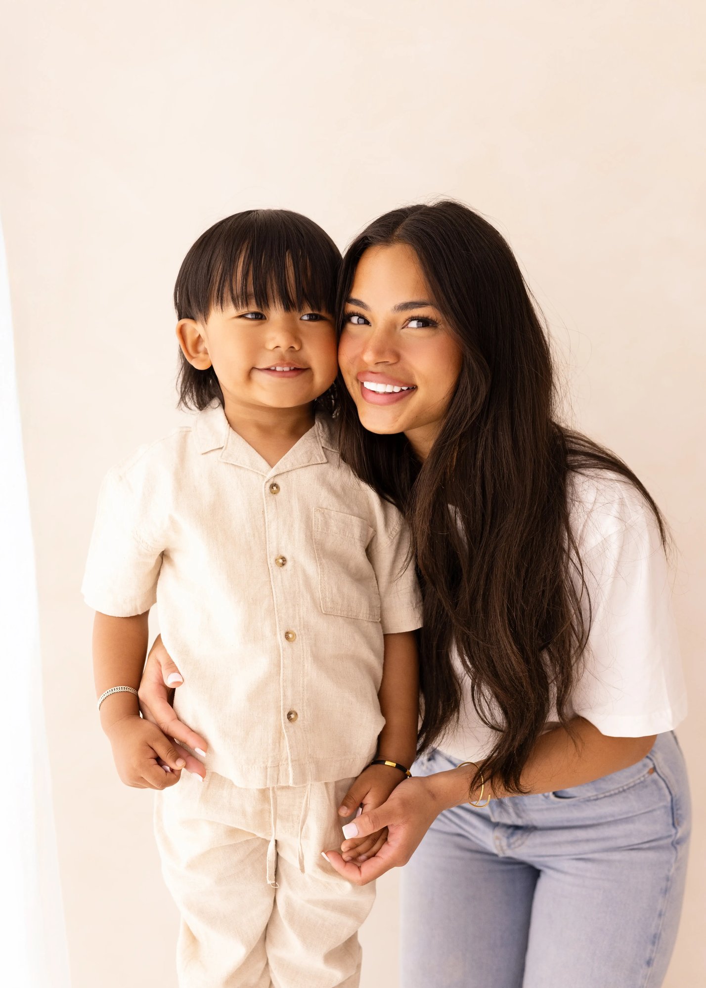 A mum and her toddler smile directly at the camera while standing close together in light-toned clothing, posed in front of a pale backdrop. A mum and her toddler smile directly at the camera while standing close together in light-toned clothing, posed in front of a pale backdrop.