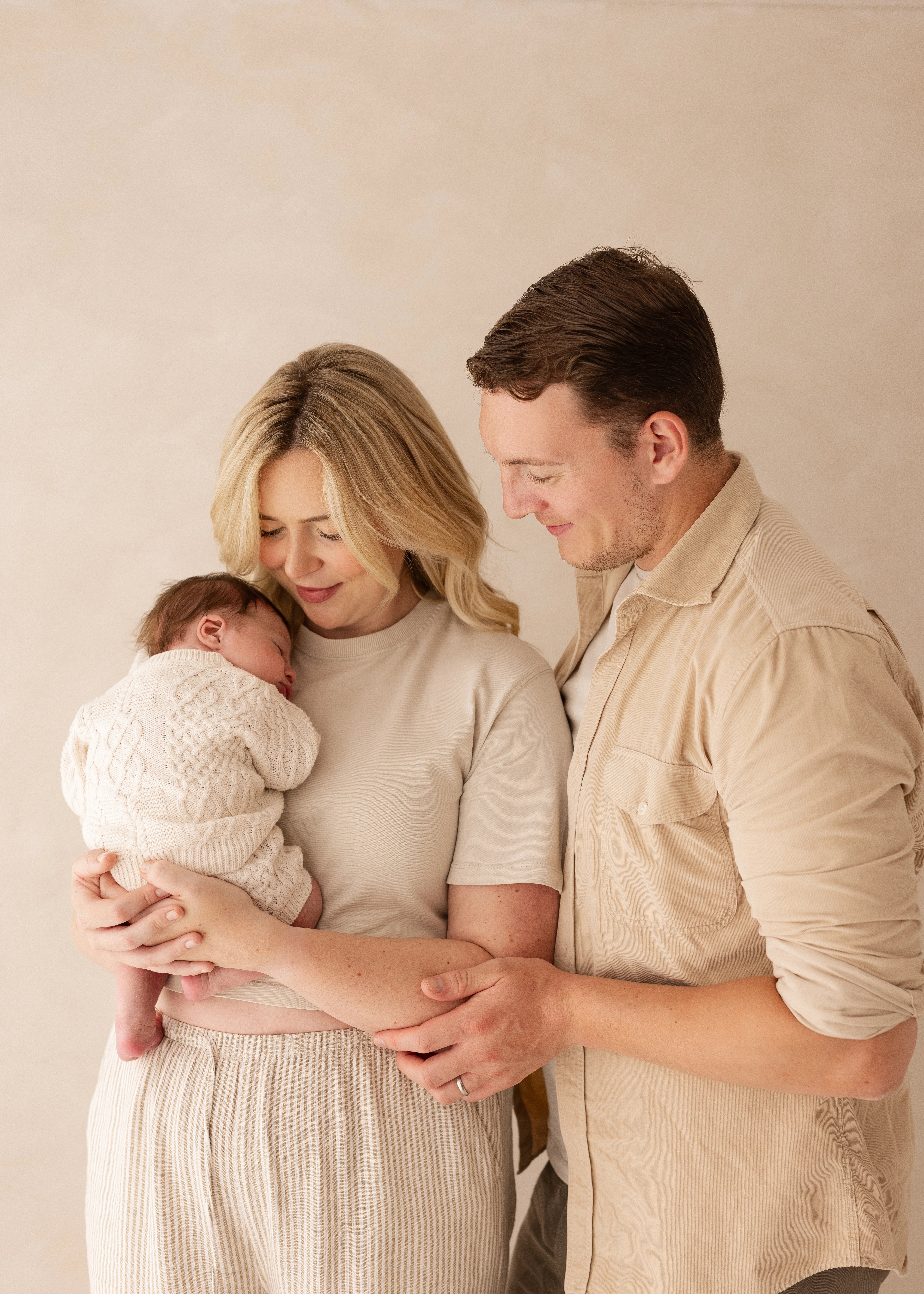 Parents gently holding their sleeping newborn in a soft, neutral-toned studio setting, captured in a warm and natural family portrait.