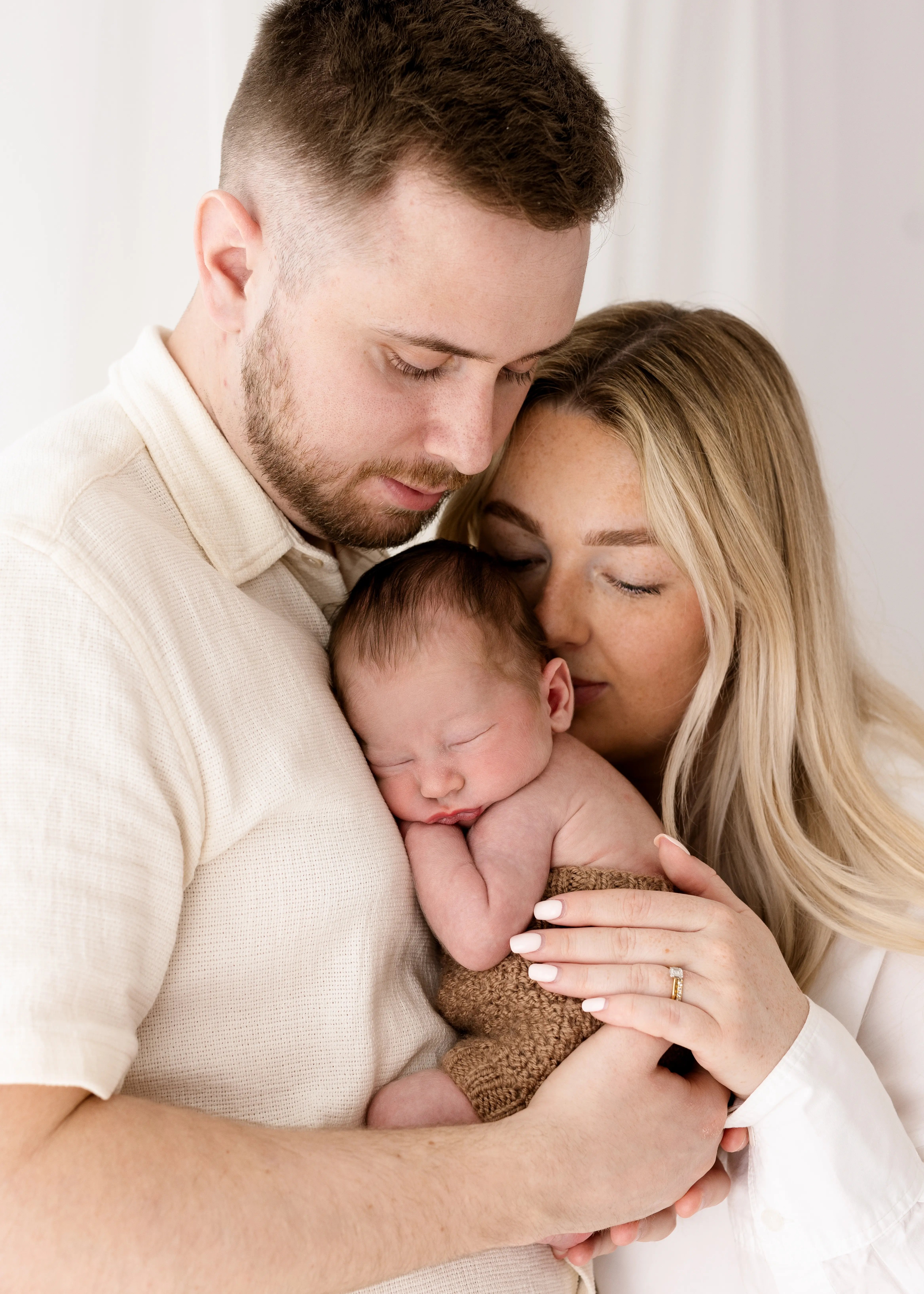 A newborn baby sleeping peacefully in their parents’ arms, with both parents gently holding and embracing their baby during a natural light newborn photoshoot.