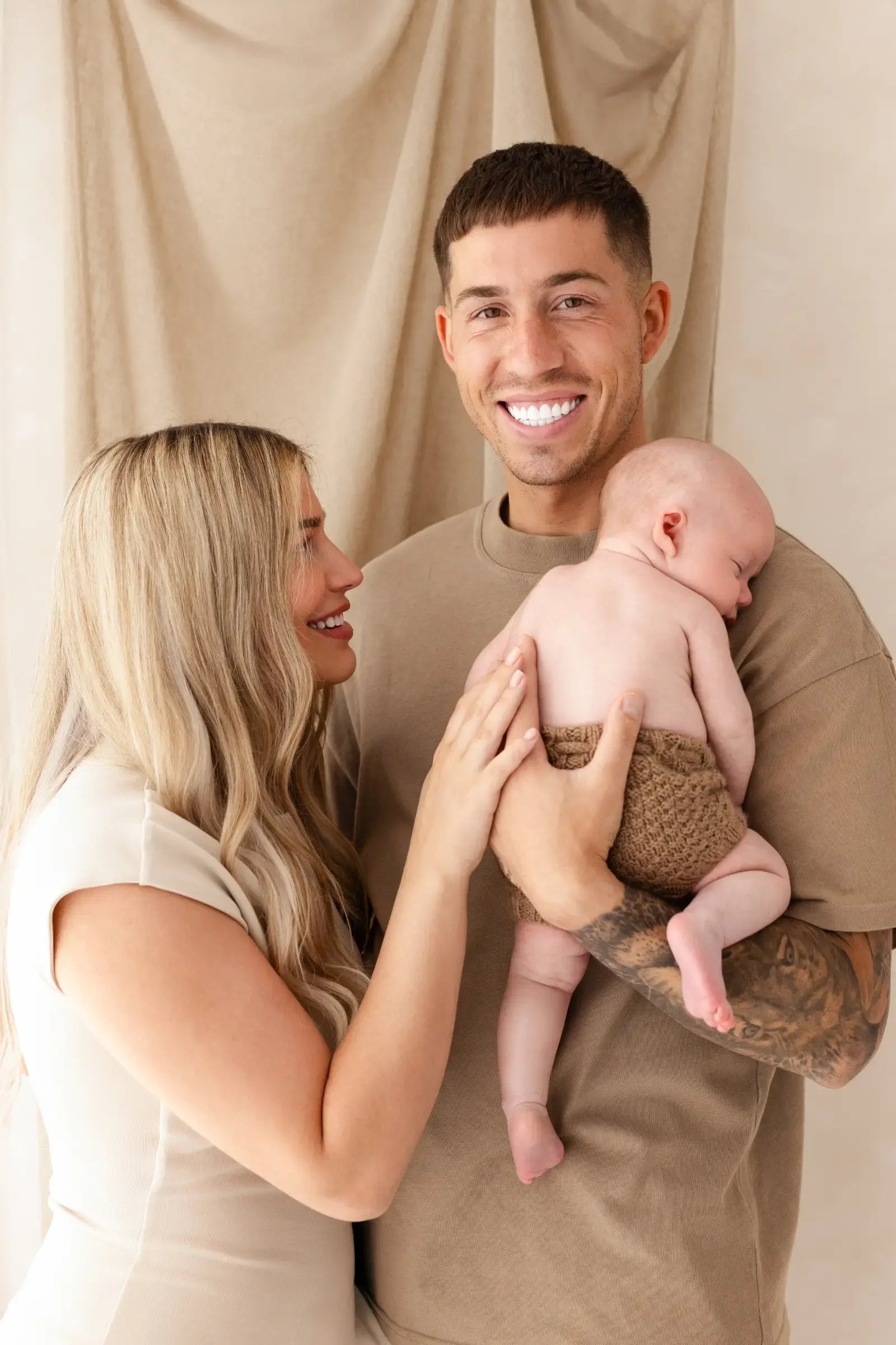 Smiling parents dressed in neutrals holding their newborn baby close in a calm and intimate family portrait, taken during a newborn photography session in Kent.
