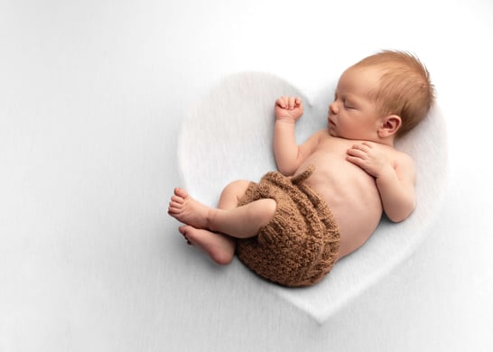 photograph of a newborn baby posed in a white fluffy heart wearing brown shorts, asleep and peaceful