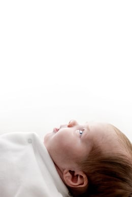 Newborn baby swaddled in white, gazing upwards in a bright, minimalistic studio setting, capturing an ethereal and peaceful first portrait.