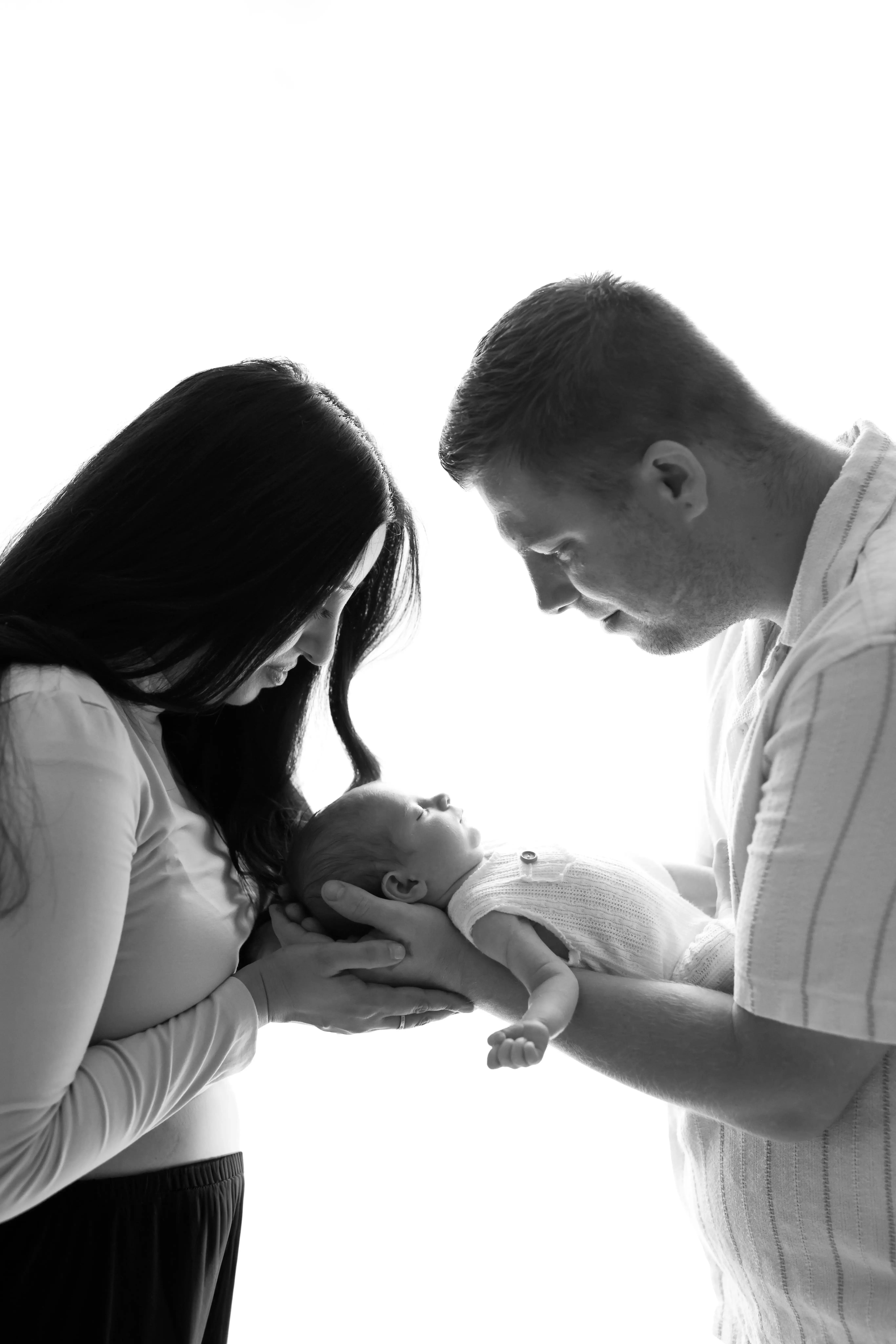 Artistic monochrome photo of parents lifting their baby, creating a striking silhouette against a light background. Conveys an intimate and timeless family moment.