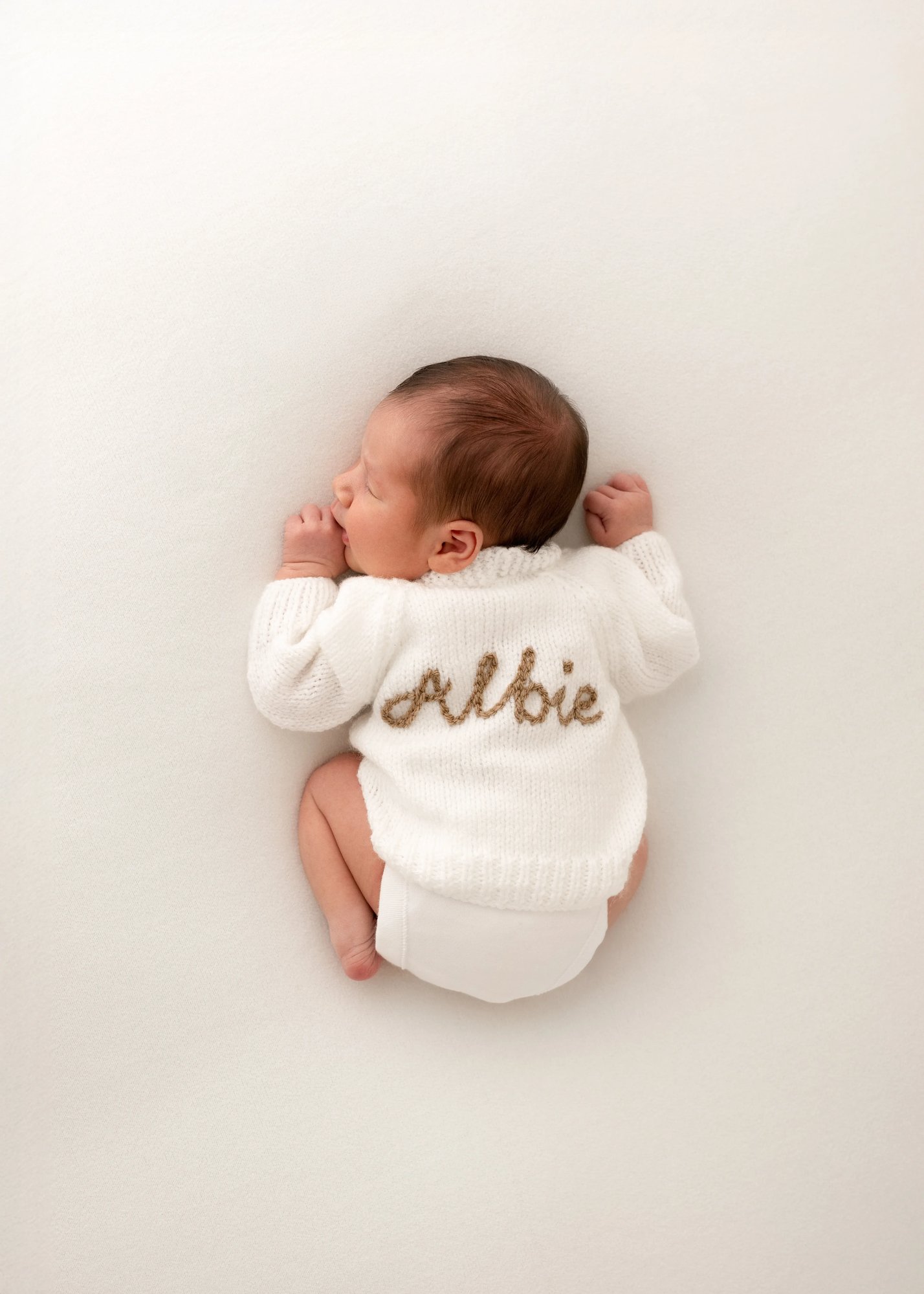 Newborn baby sleeping on a cream blanket, wearing a soft white knitted jumper embroidered with their name on the back, photographed in a calm minimalist studio setup.