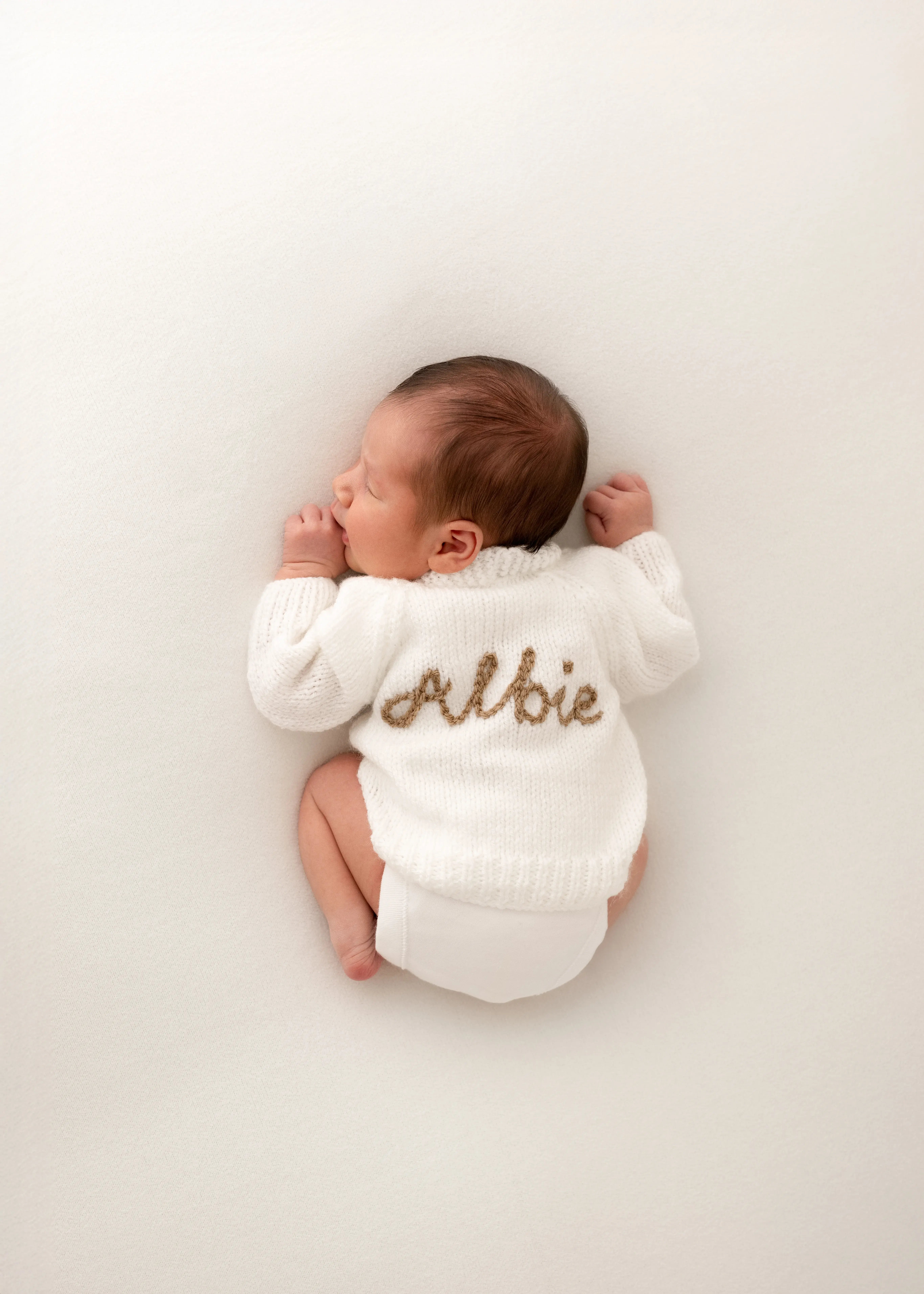Newborn baby sleeping on a cream blanket, wearing a soft white knitted jumper embroidered with their name on the back, photographed in a calm minimalist studio setup.