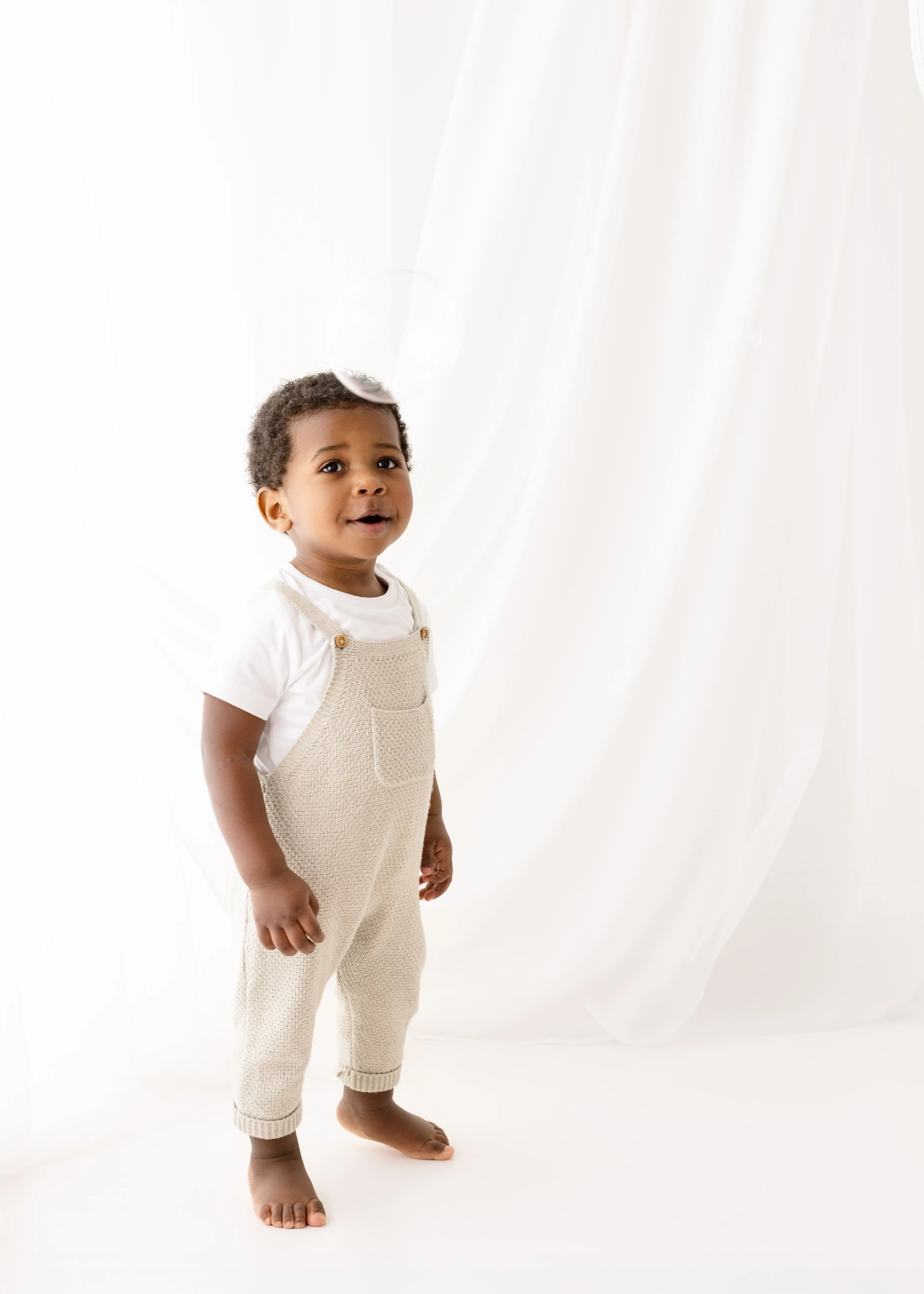 Playful toddler boy standing barefoot in a white studio, wearing a cream knitted romper and white t-shirt, gazing curiously at a floating bubble above his head.