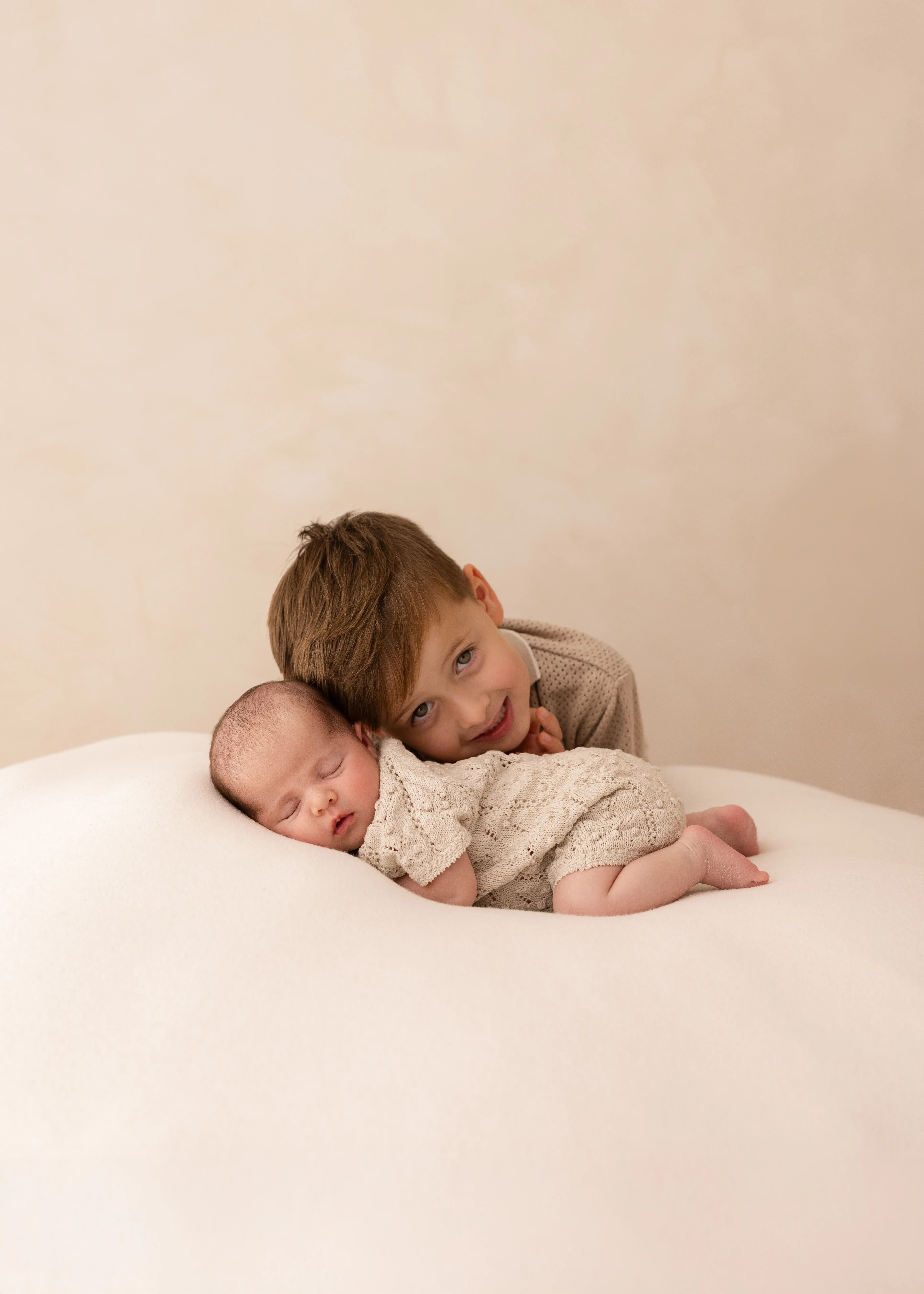 Older brother smiling as he gently cuddles his sleeping newborn sibling on a soft cream blanket during a natural newborn photoshoot in Kent.