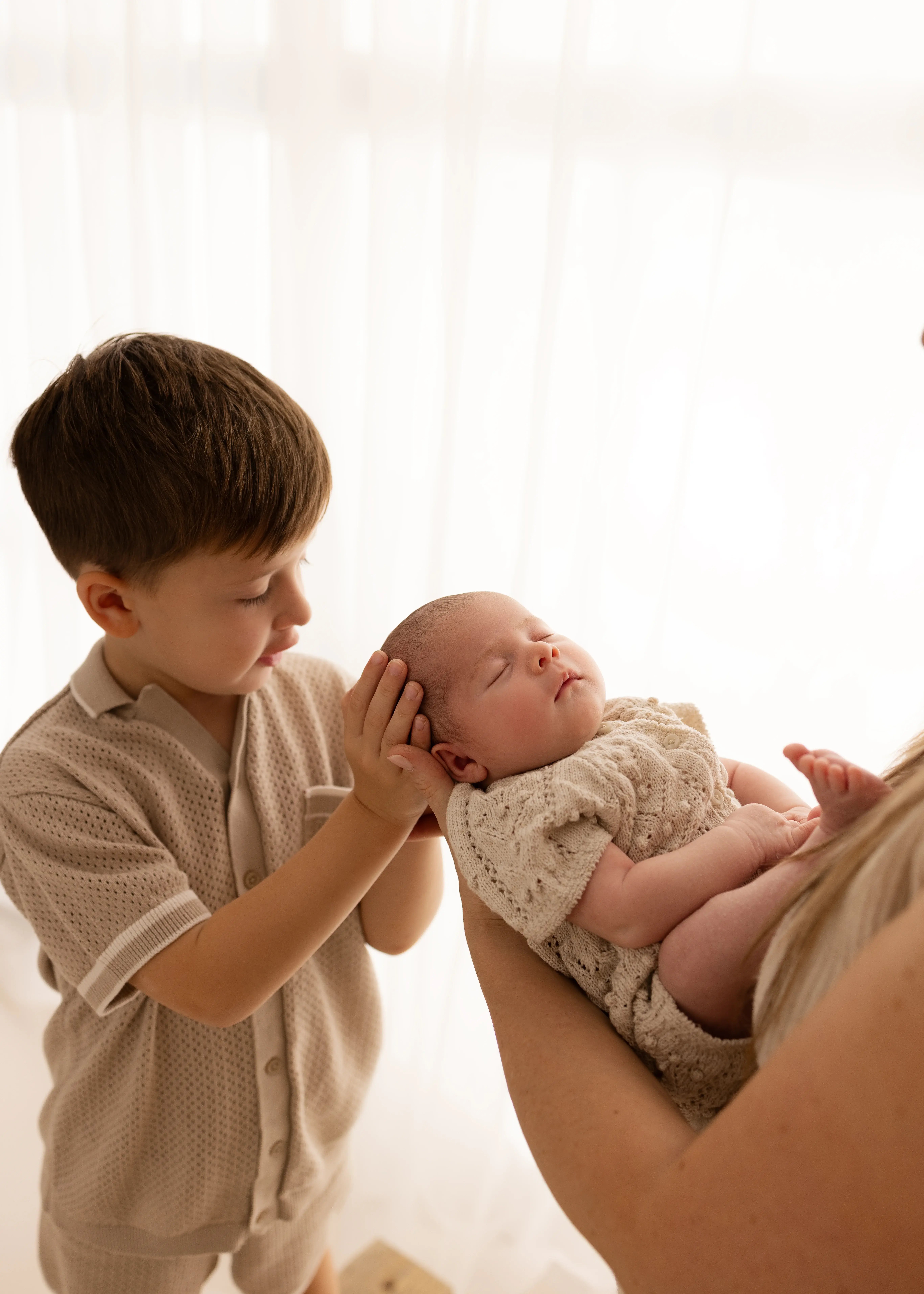 Older brother gently holding his newborn sibling’s head while the baby sleeps in a parent’s arms during a natural newborn photoshoot in Kent.
