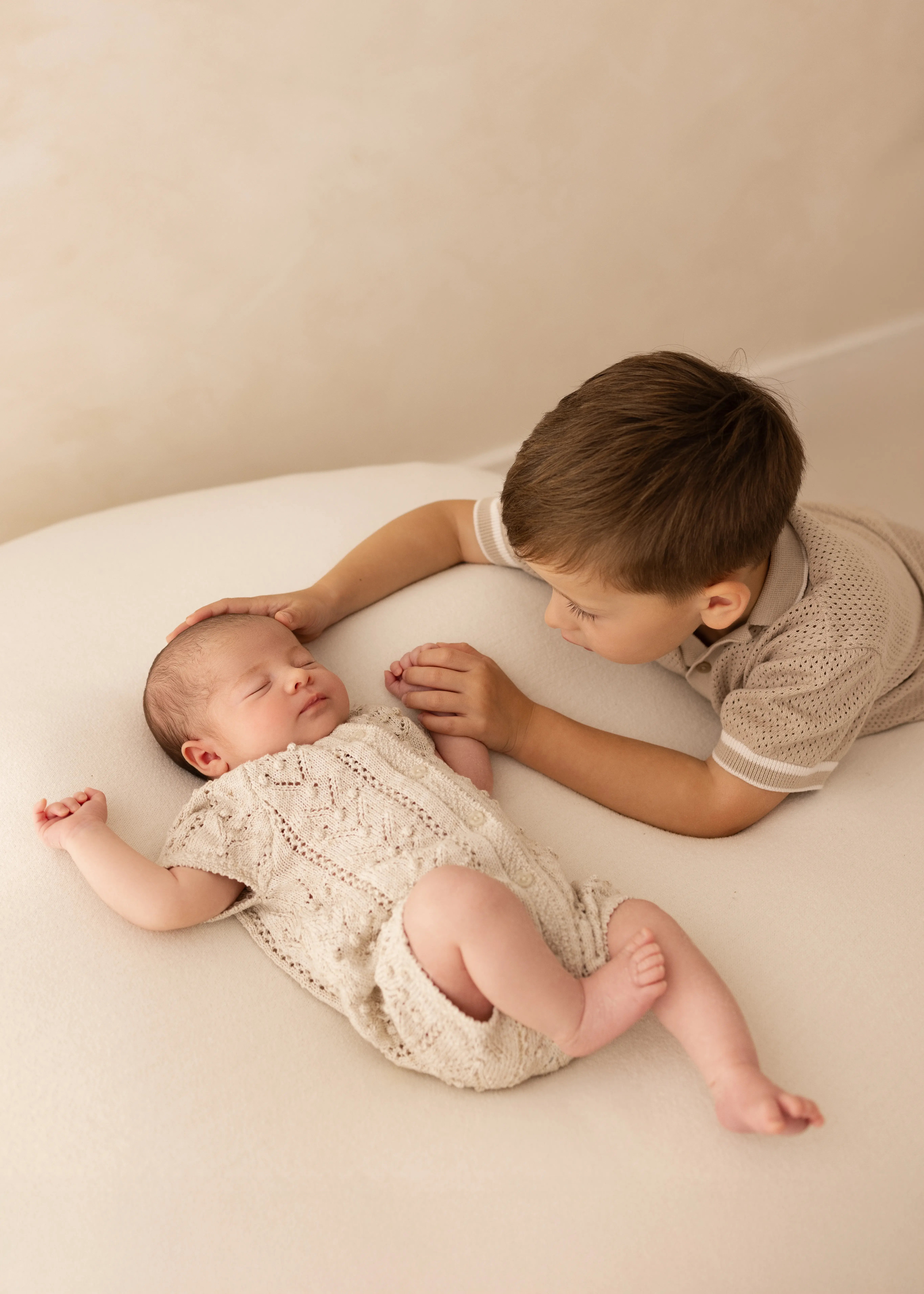 Older brother lying beside his newborn sibling, gently touching her head and holding her hand during a soft, natural newborn photoshoot in Kent.