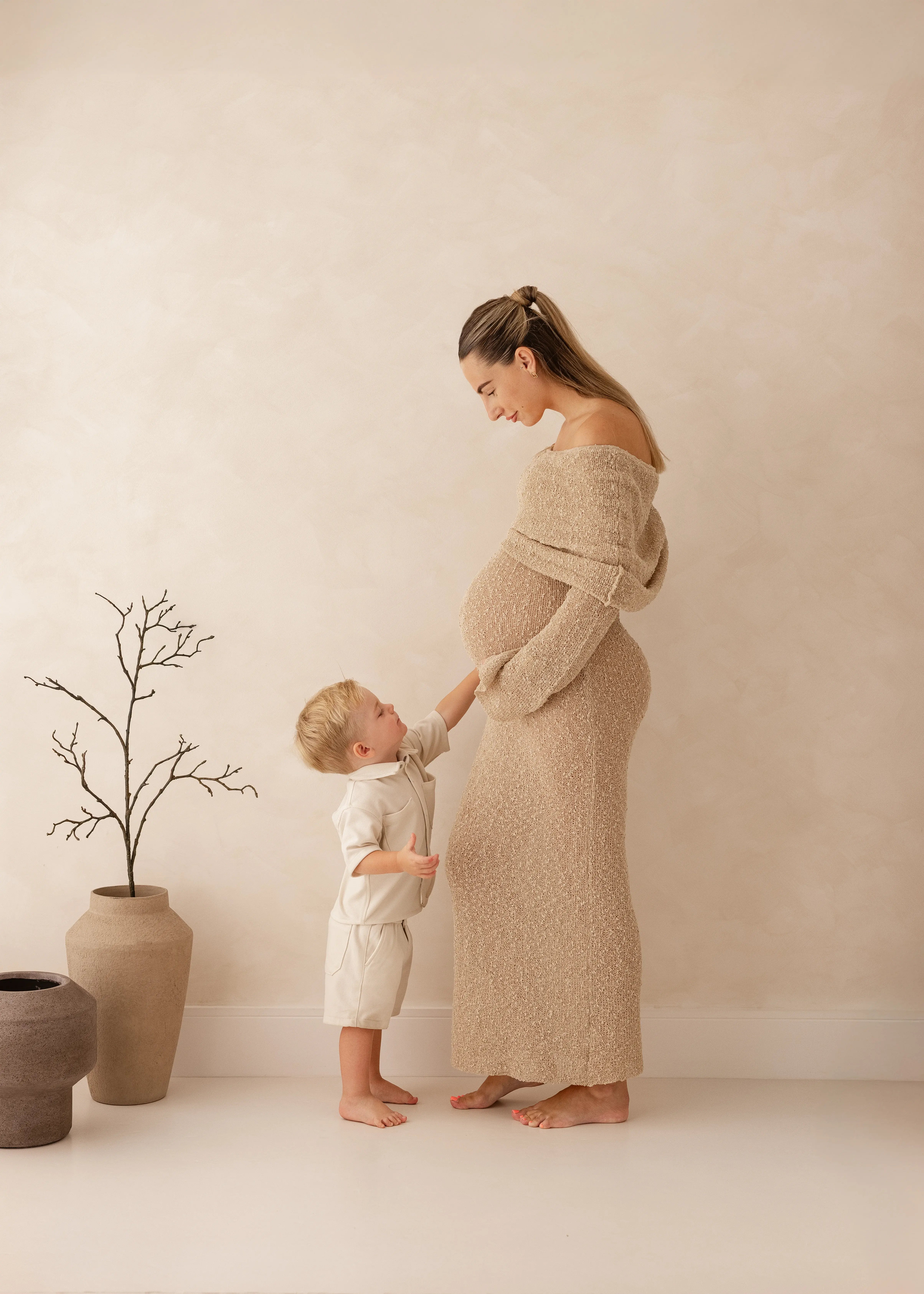 Pregnant mother standing in profile while her toddler reaches up to touch her baby bump during a soft, neutral maternity photoshoot in Kent.