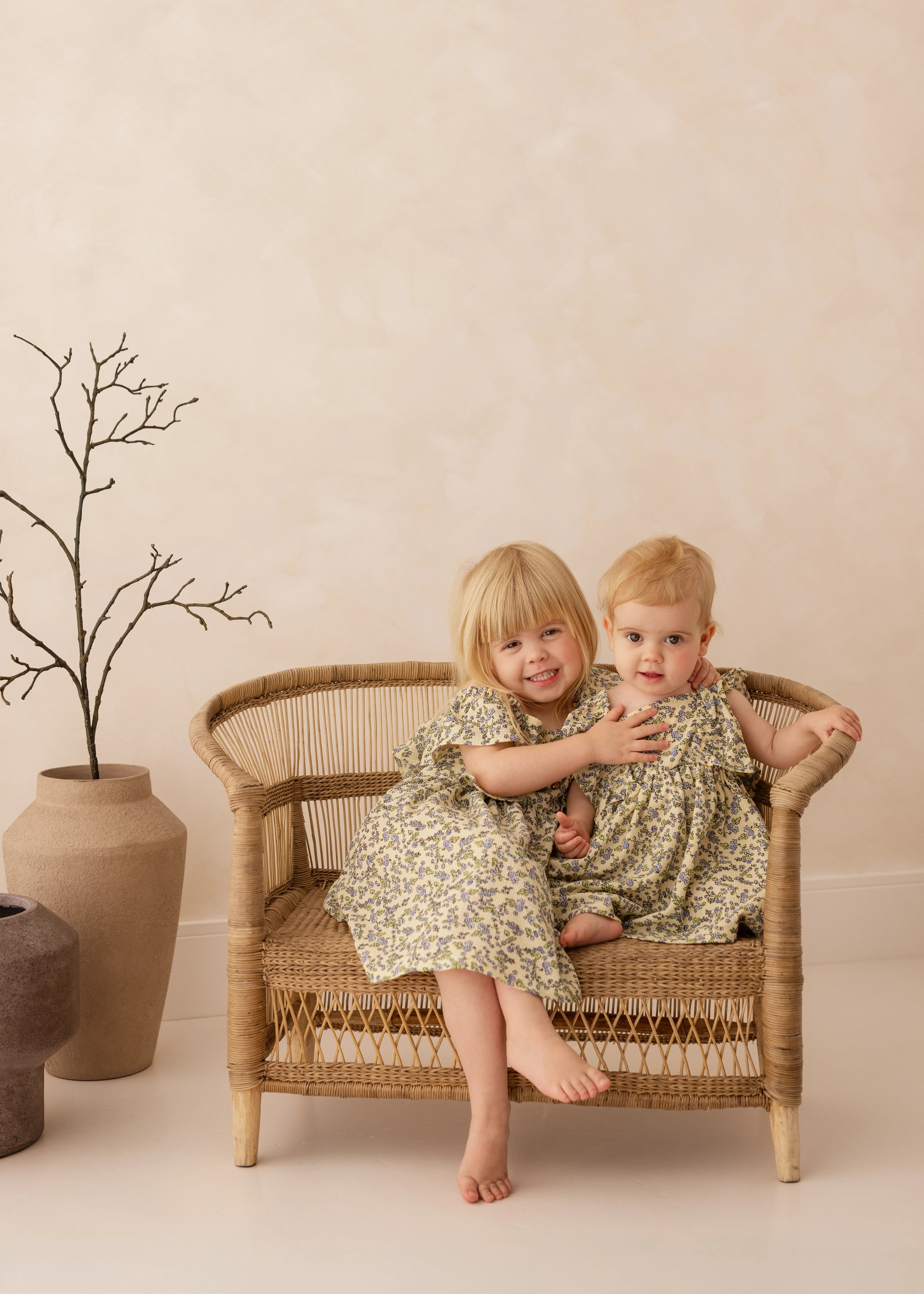 Two young sisters in matching floral dresses sitting on a wicker bench, smiling together during a natural family studio photography session.