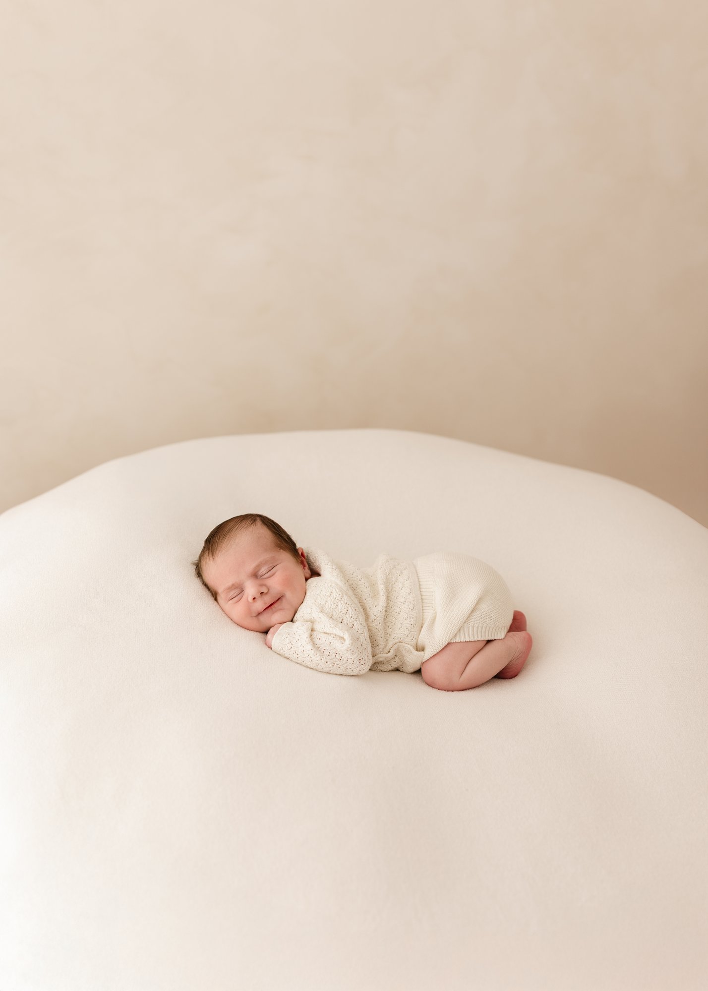 Newborn baby sleeping on a soft cream blanket, smiling gently while wearing a knitted cream outfit during a natural newborn photography session in Kent.