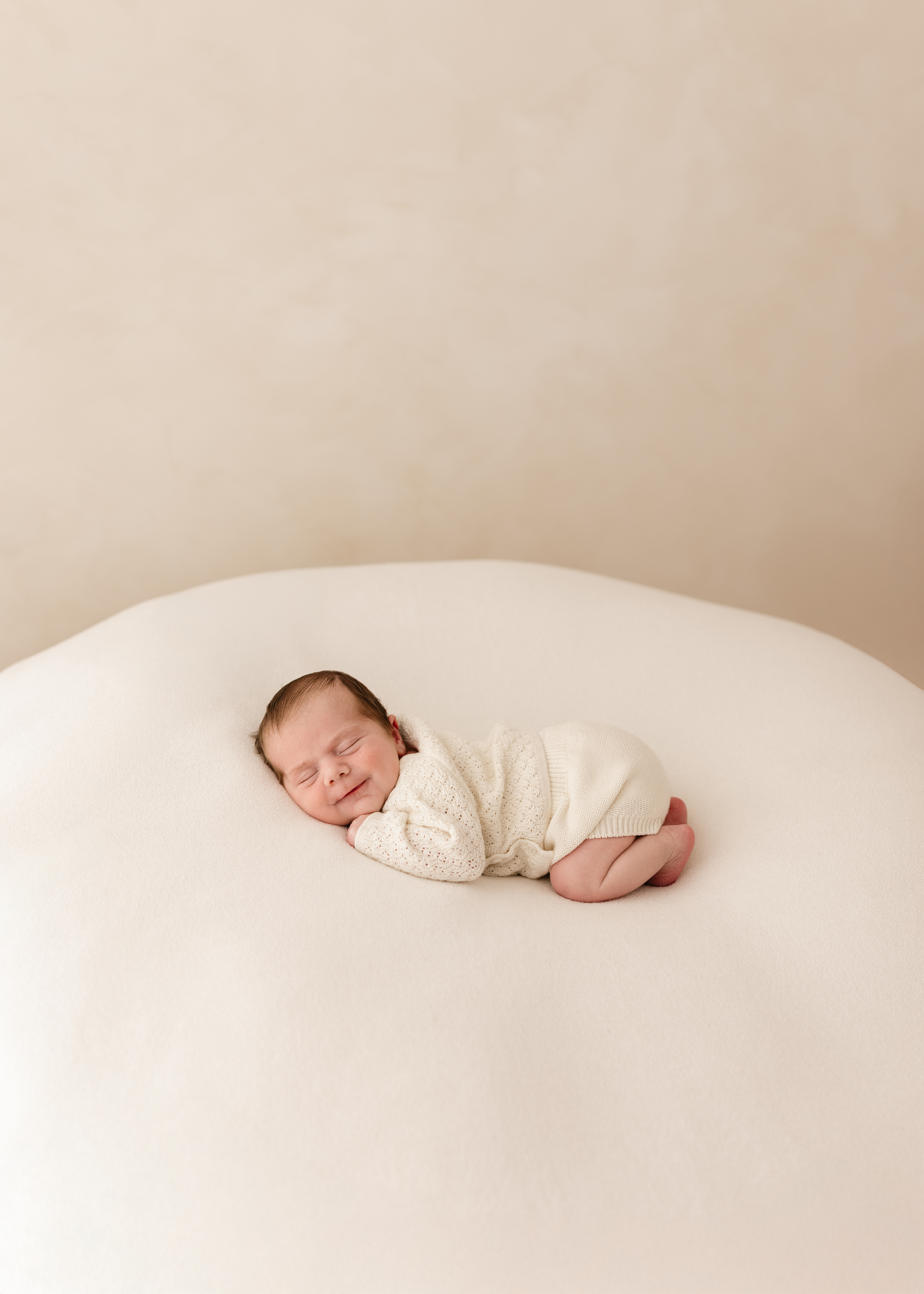Newborn baby sleeping on a soft cream blanket, smiling gently while wearing a knitted cream outfit during a natural newborn photography session in Kent.